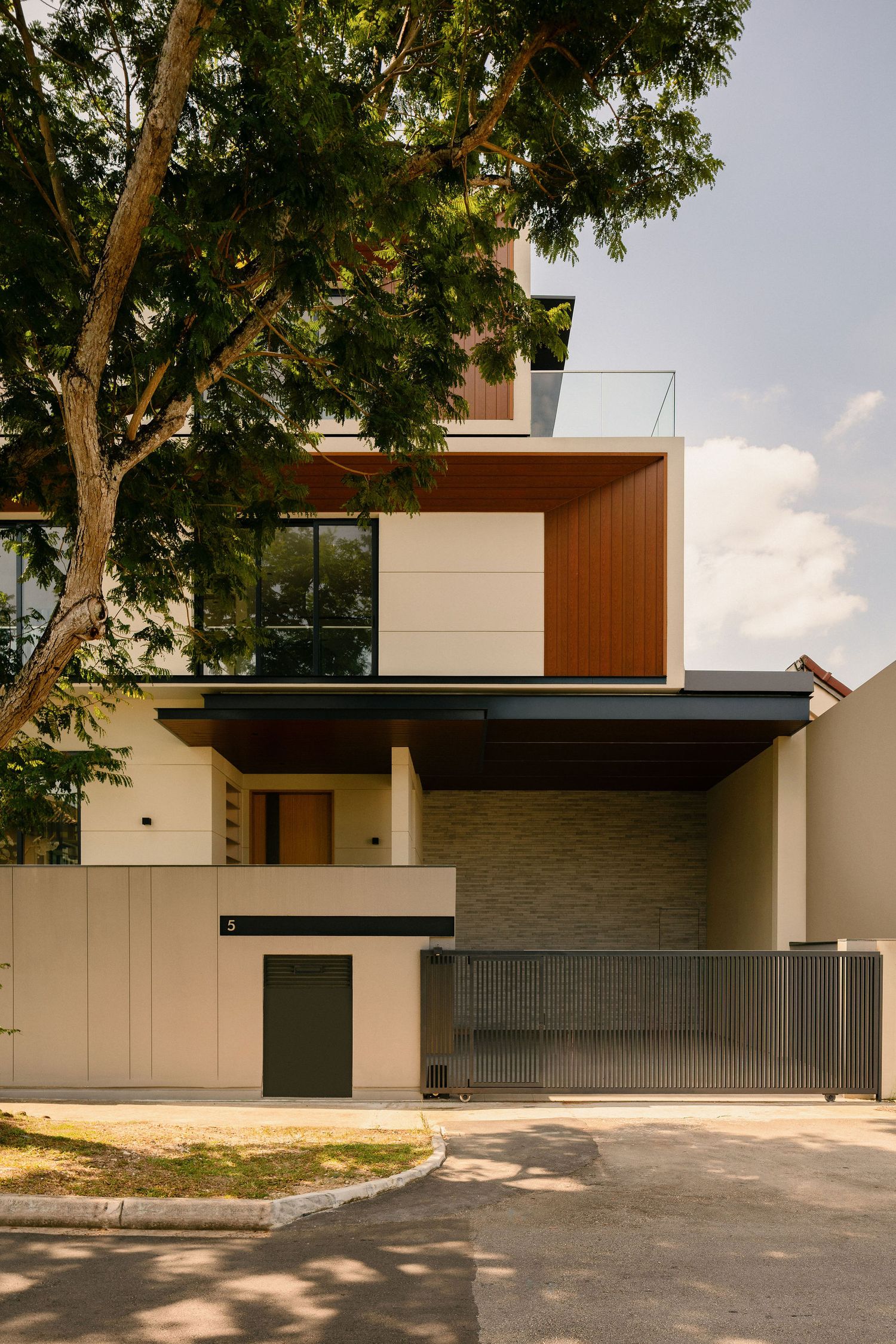 Modern two-story house with wood-paneled upper facade, glass balcony, stone accent wall, and sliding gate, framed by a large tree.