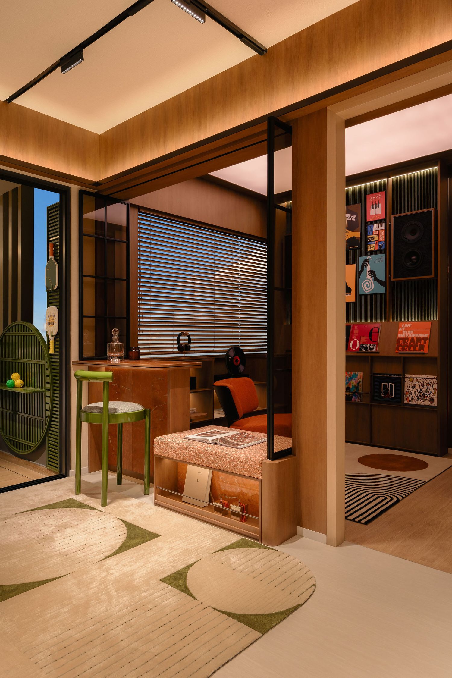 Warm wood-paneled home study with a built-in desk, green stool, window blinds, vinyl record shelves and a geometric area rug.