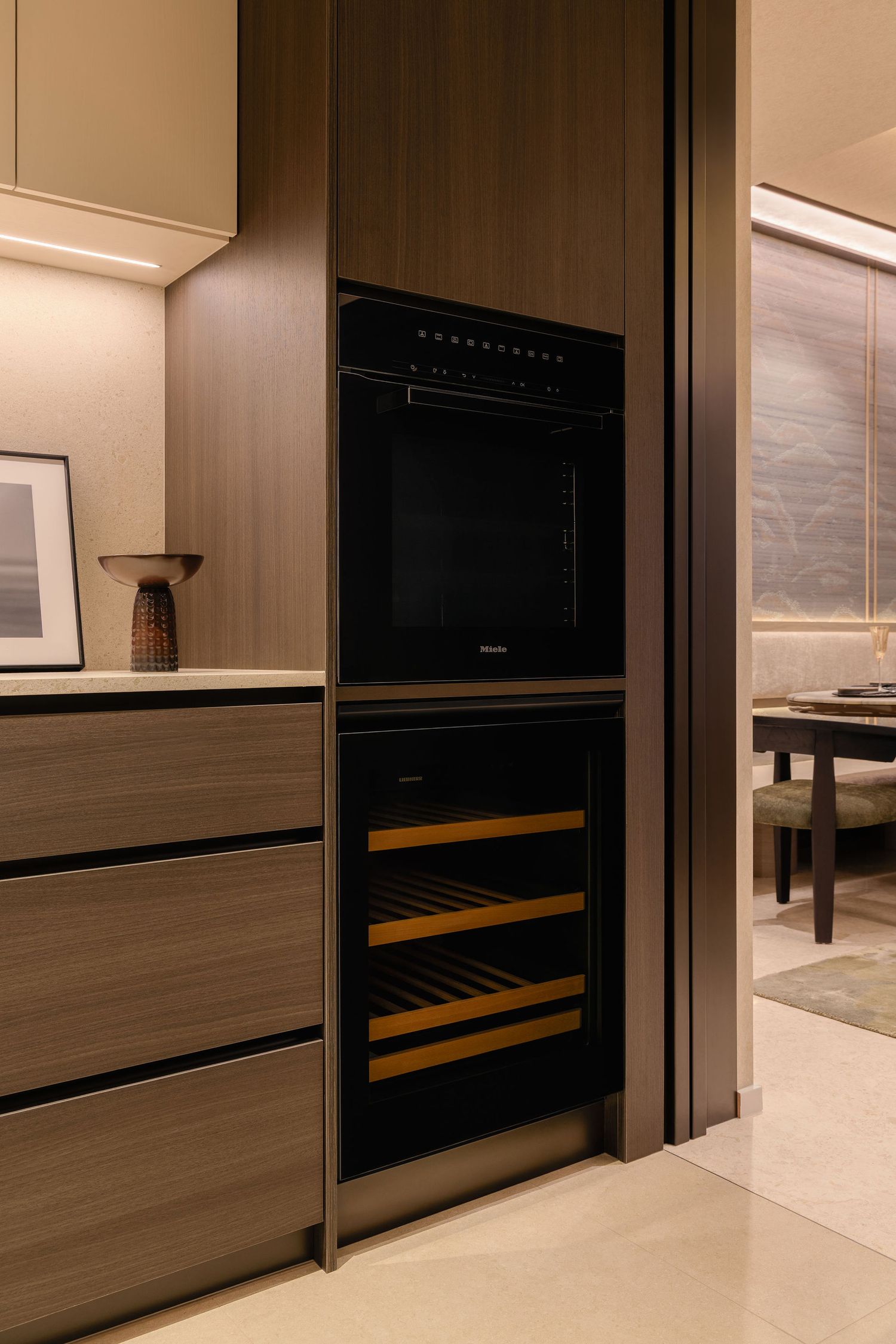 Modern kitchen with a built-in black oven above a wine cooler with wooden shelves, set in warm wood cabinetry near a dining area.