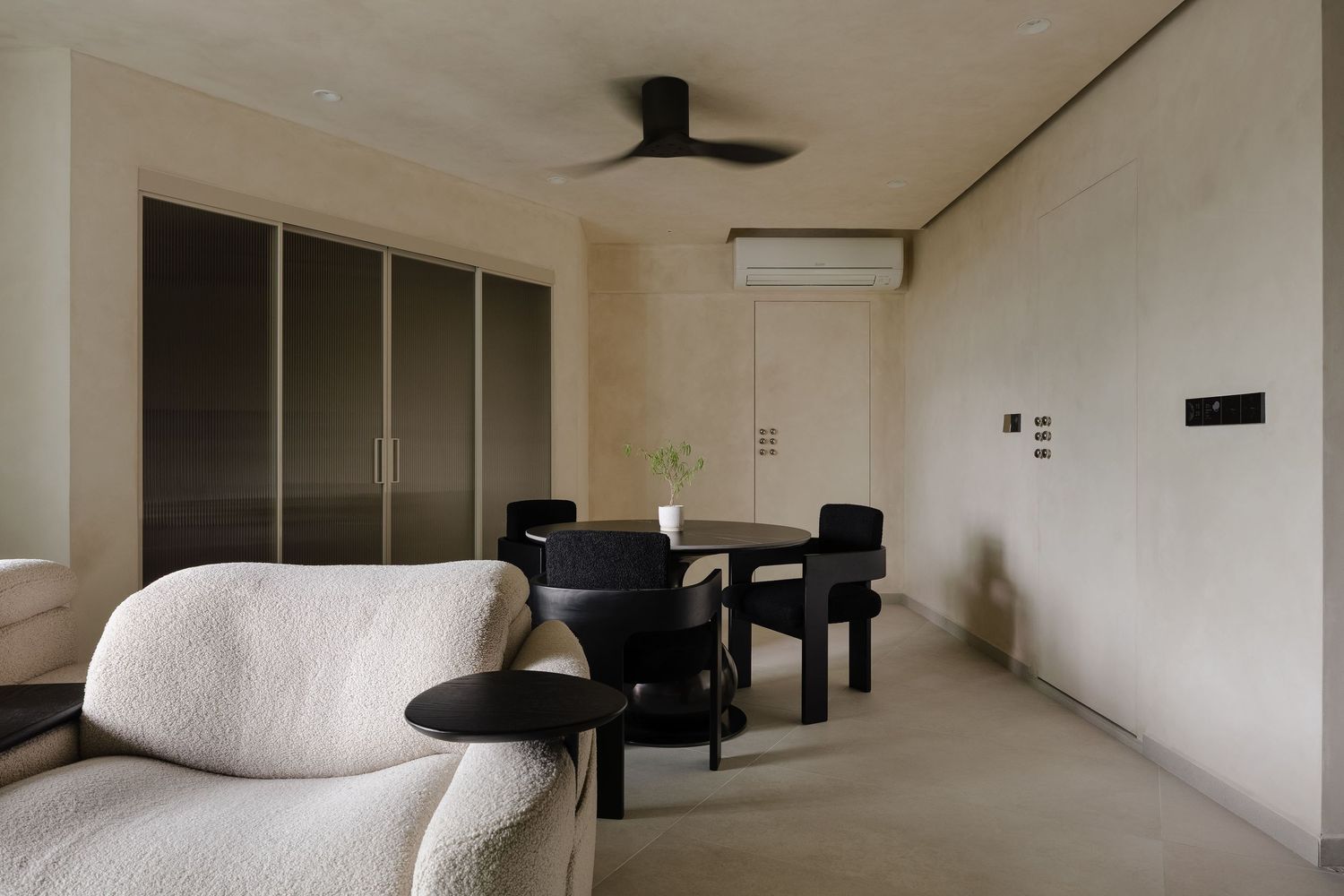 Minimalist living room with white boucle sofa, black dining set, ceiling fan, and beige walls in a modern apartment.