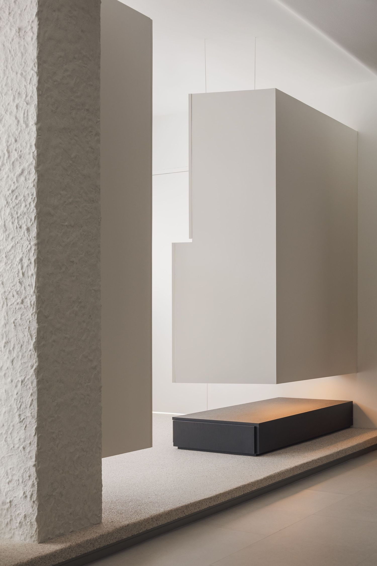 Minimalist interior with textured stone column, geometric beige wall panels, and a sleek dark rectangular object on a raised platform.