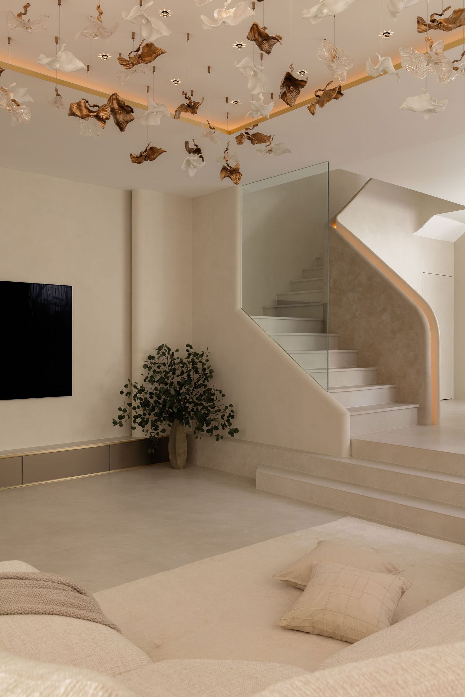 Modern minimalist living room with beige tones, decorative ceiling installation, glass staircase, TV wall, and lush green plant.