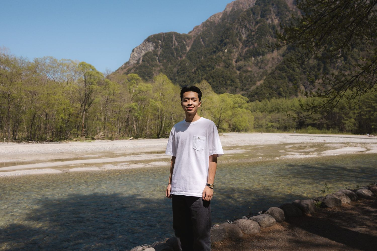 Young man in white t-shirt standing by a clear mountain river with lush green trees and rocky peaks behind him.