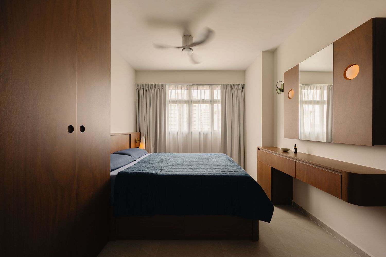Modern bedroom with dark wood wardrobe, blue bedding, ceiling fan, sheer curtains, and a floating wooden vanity with mirror.