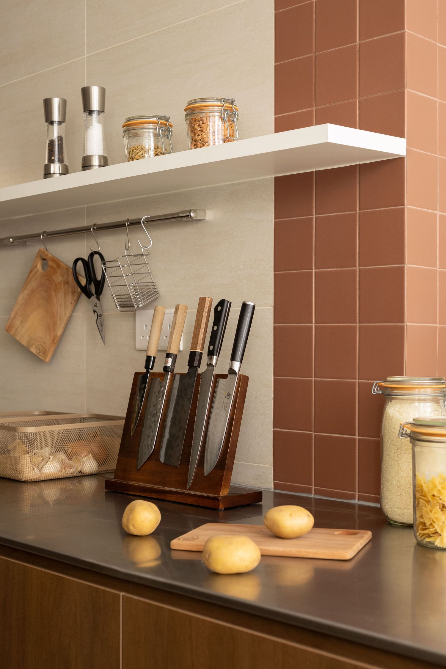 Modern kitchen counter with wooden knife block, floating shelf, glass storage jars and fresh buns on dark countertop.
