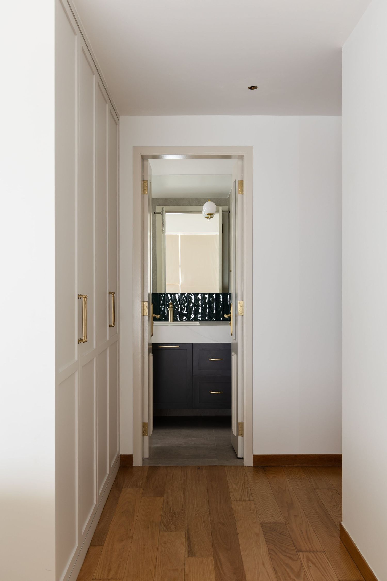 Minimalist hallway with hardwood floors leads to modern black vanity bathroom through white doorframe.