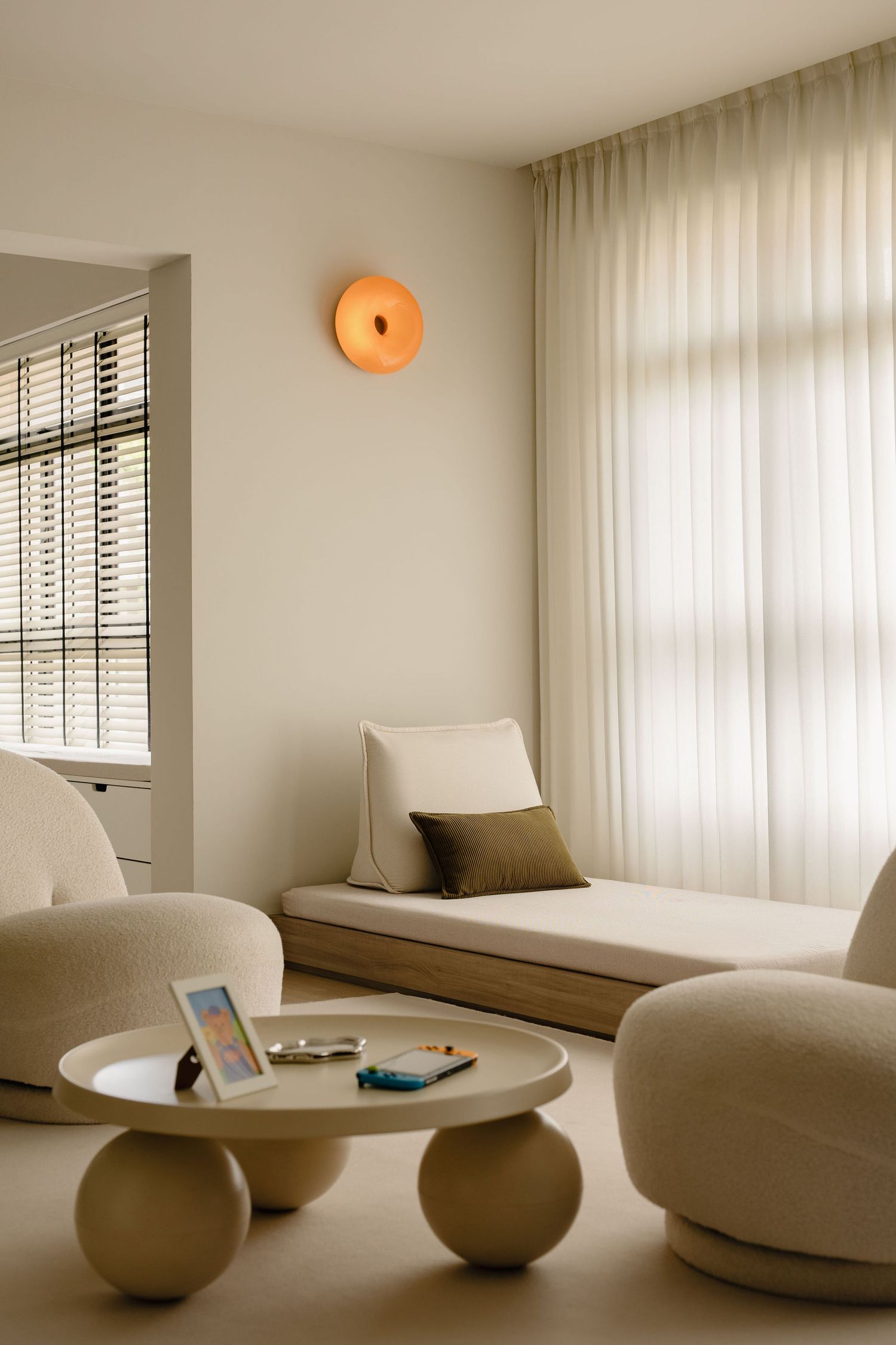 Minimalist living room with cream bouclé chairs, round coffee table, daybed, sheer curtains, and warm orange wall sconce.