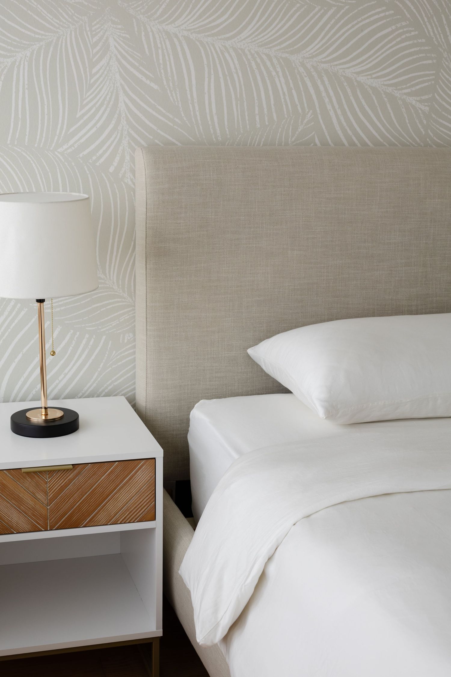 Modern minimalist hotel bed with crisp white linens and beige upholstered headboard against a textured wall.