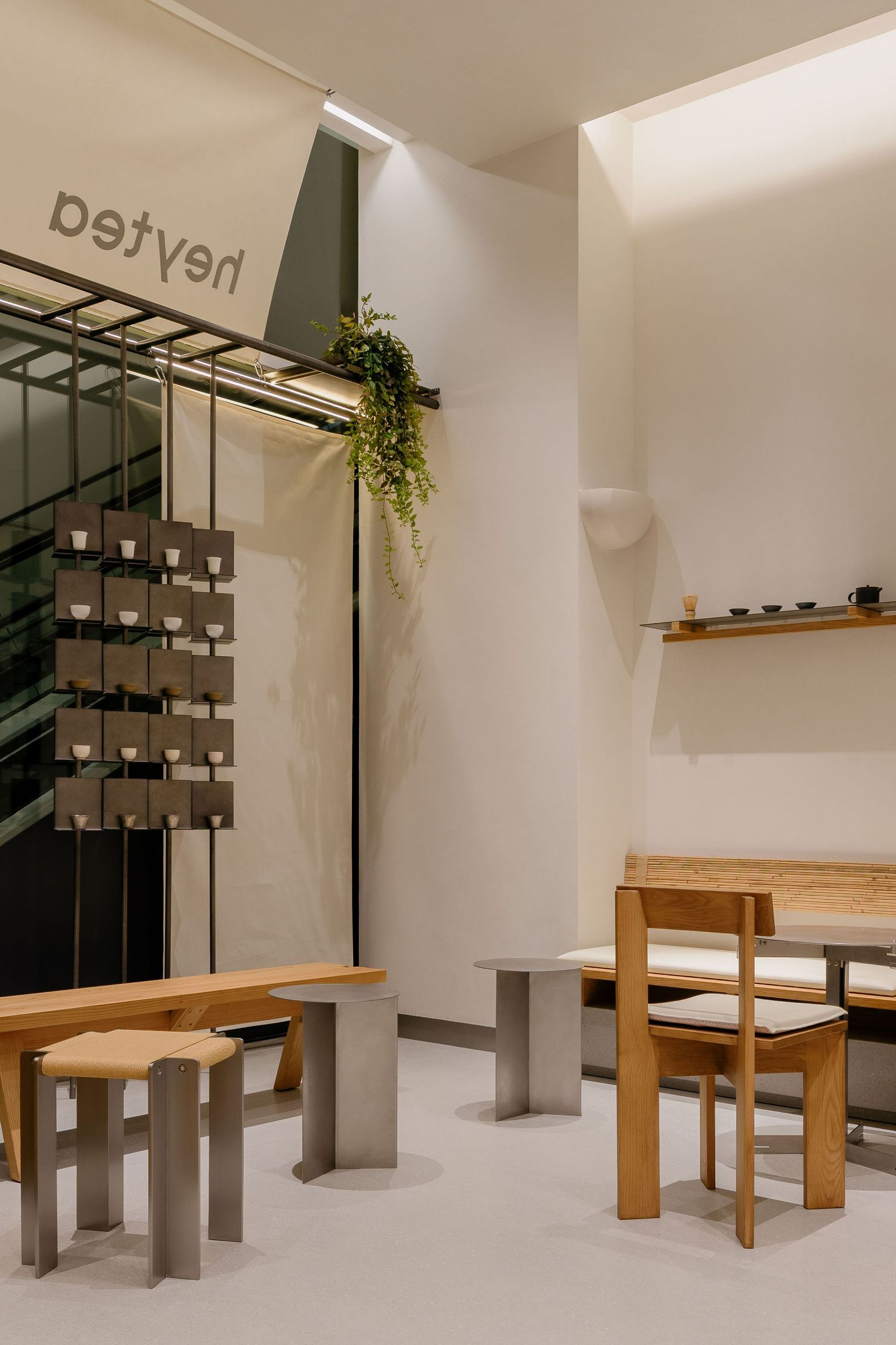Minimalist cafe interior with wooden chairs, metal stools, a trailing plant, a shelf of ceramics and a Heyteya banner on white walls.