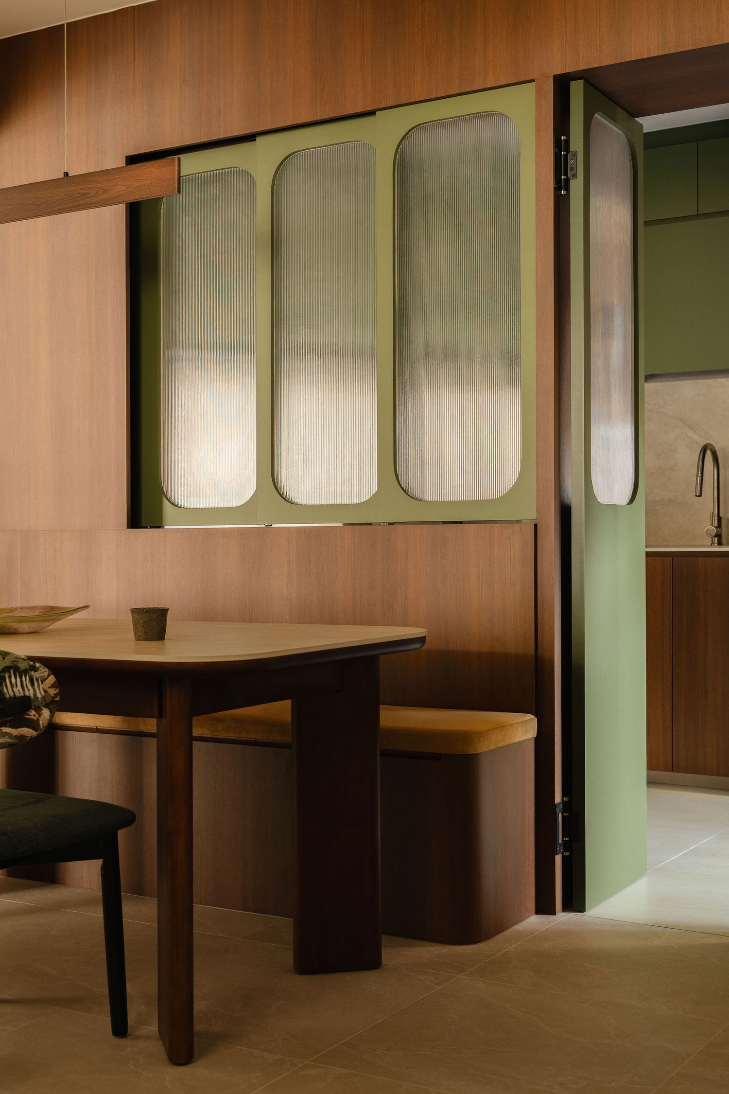 Walnut dining nook with a mustard banquette, wooden table and green folding doors with fluted glass panels opening to a kitchen.