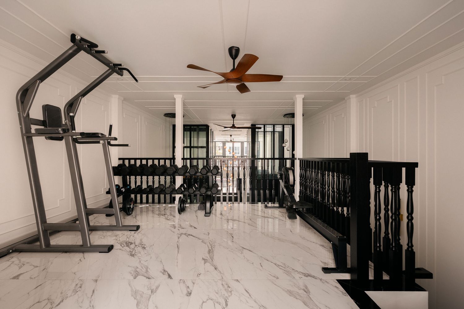 Elegant home gym with white marble flooring, a power tower, dumbbell rack, ceiling fans and a black wooden balustrade.