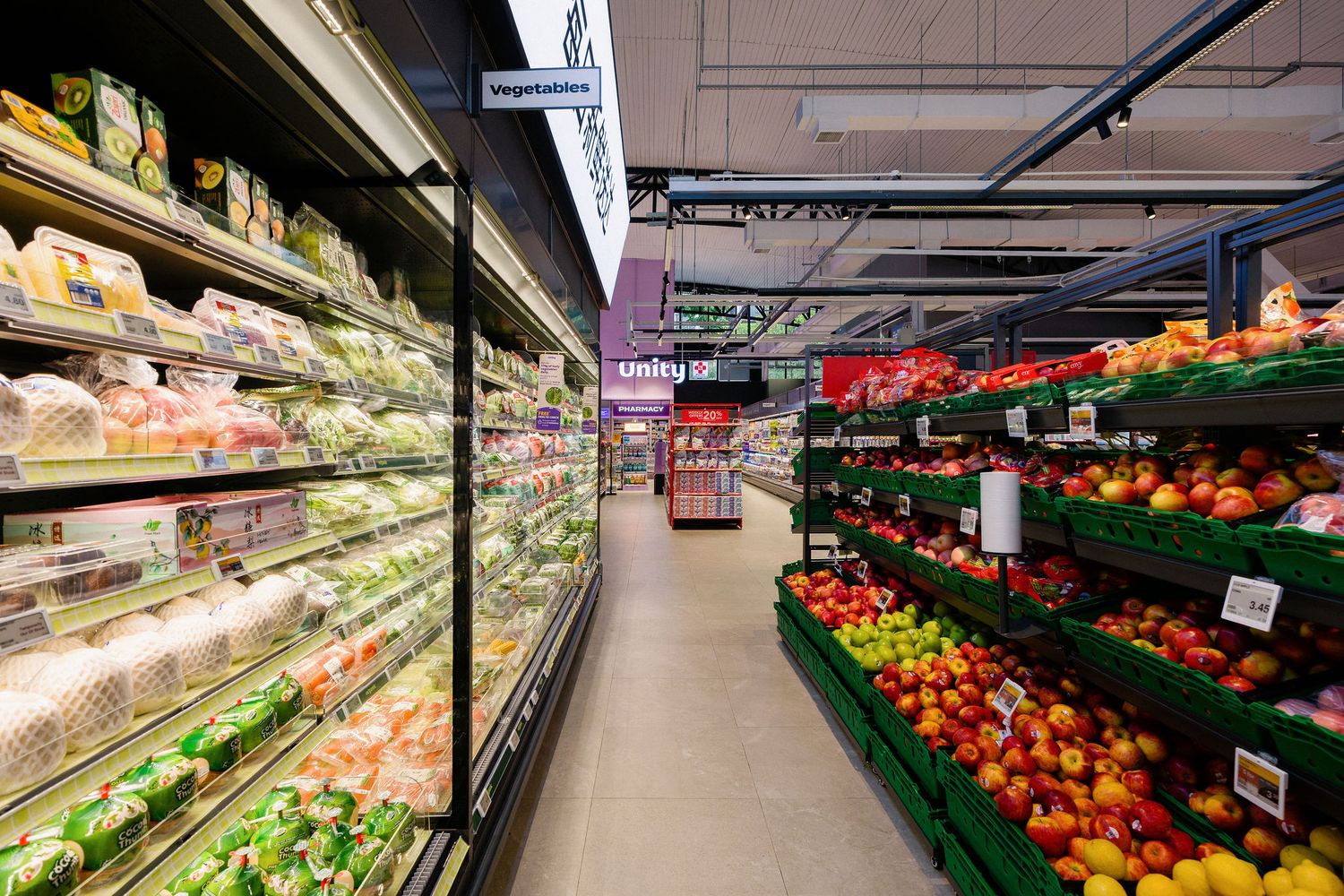 Supermarket produce aisle with refrigerated shelves of packaged goods on the left and displays of fresh red apples and fruit on the right.