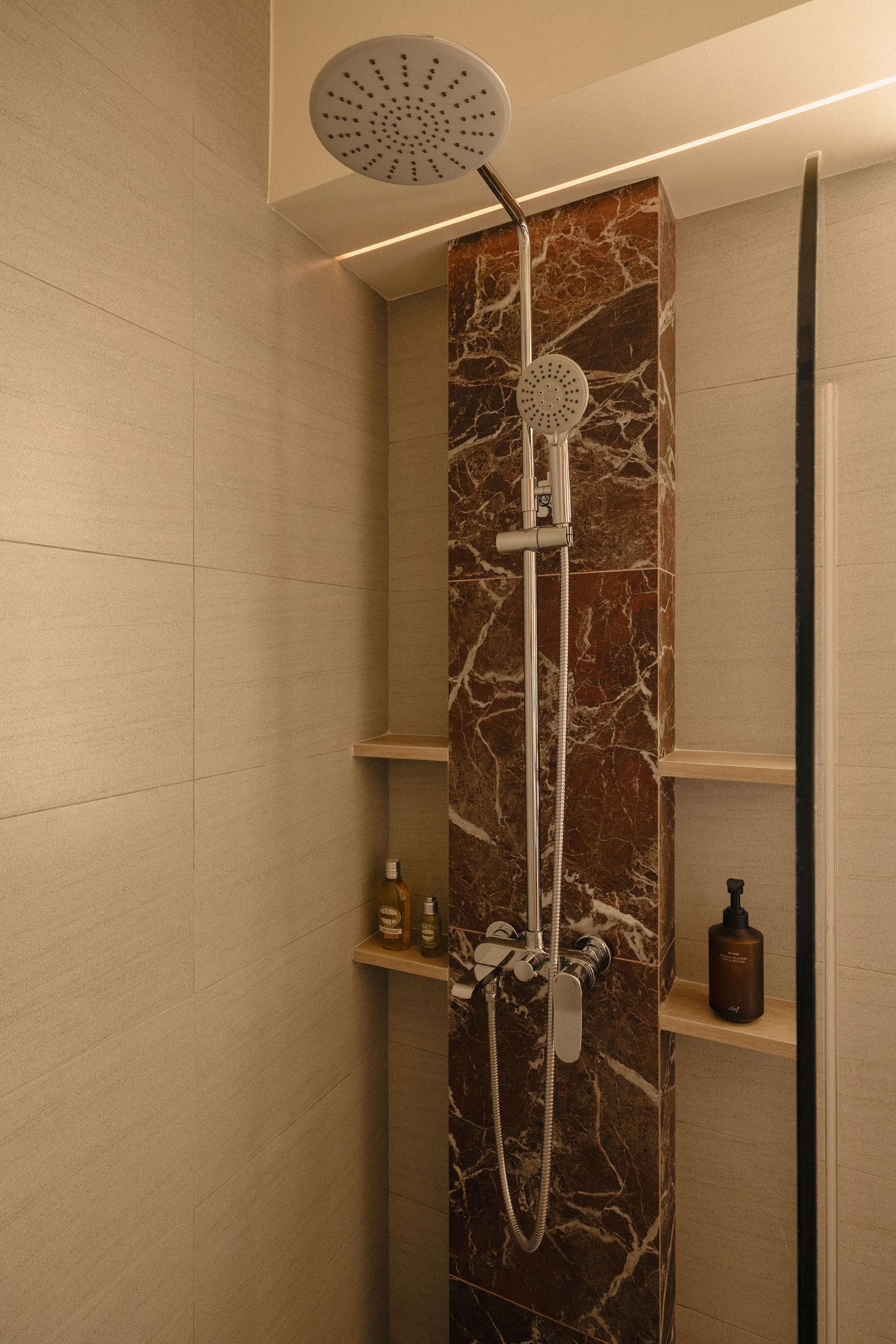 Modern shower with rain showerhead and handheld sprayer on a red marble accent column, beige tiles, and built-in wooden niche shelves.