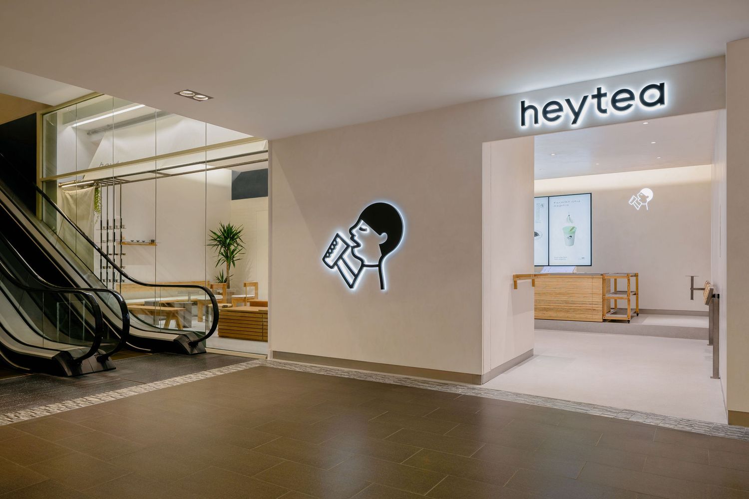 Modern Heytea storefront entrance with a glowing illuminated logo sign beside an escalator in a bright, minimalist shopping mall.