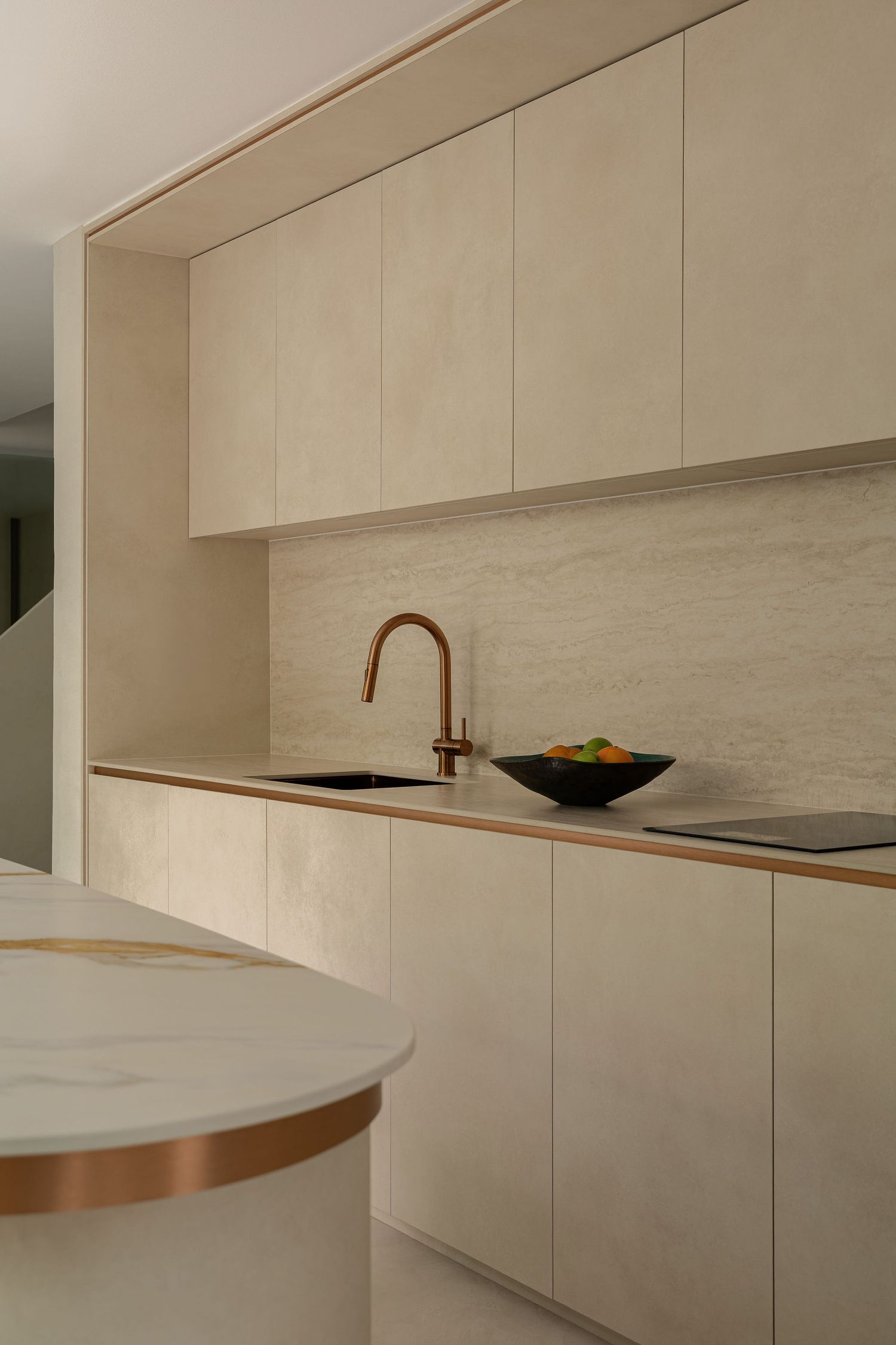 Modern minimalist kitchen with cream cabinetry, marble backsplash, brass faucet, and white marble island with copper trim.