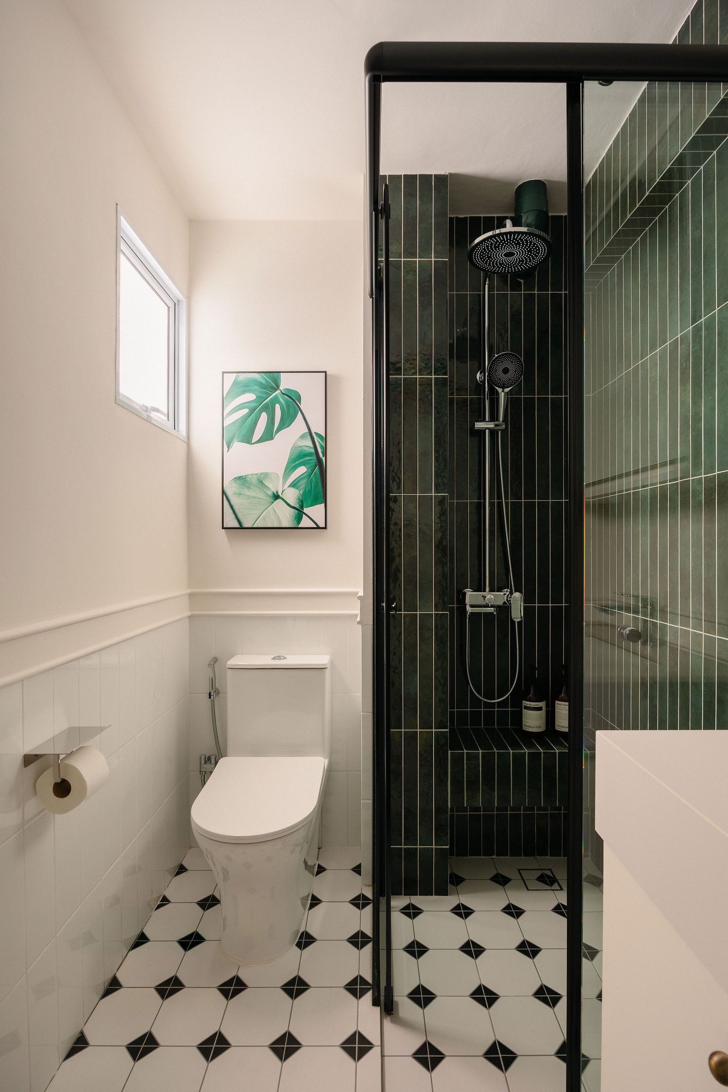 Modern bathroom with green vertical tiles, black frame shower, white toilet, monstera wall art, and classic black and white floor tiles.
