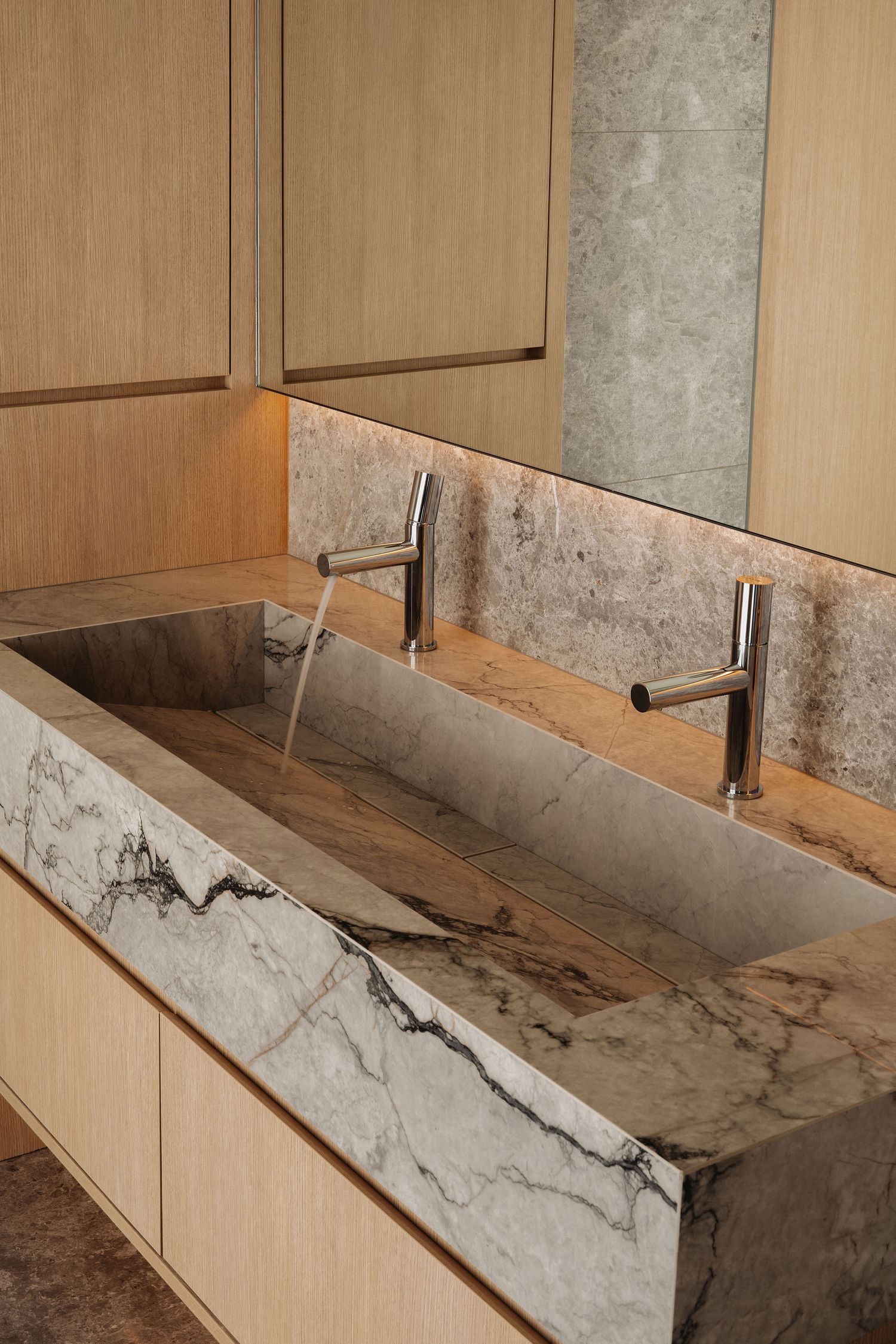 Luxury marble trough sink with dual chrome faucets, veined stone countertop, and warm wood cabinetry in modern bathroom.