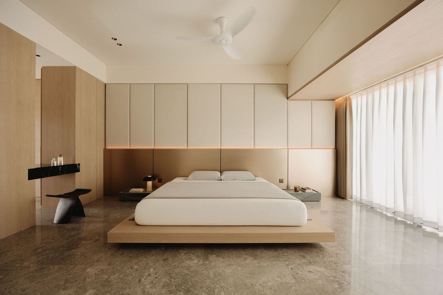 Minimalist bedroom with low platform bed, light wood cabinetry, concrete floors, white linens, and sheer curtains in neutral tones.