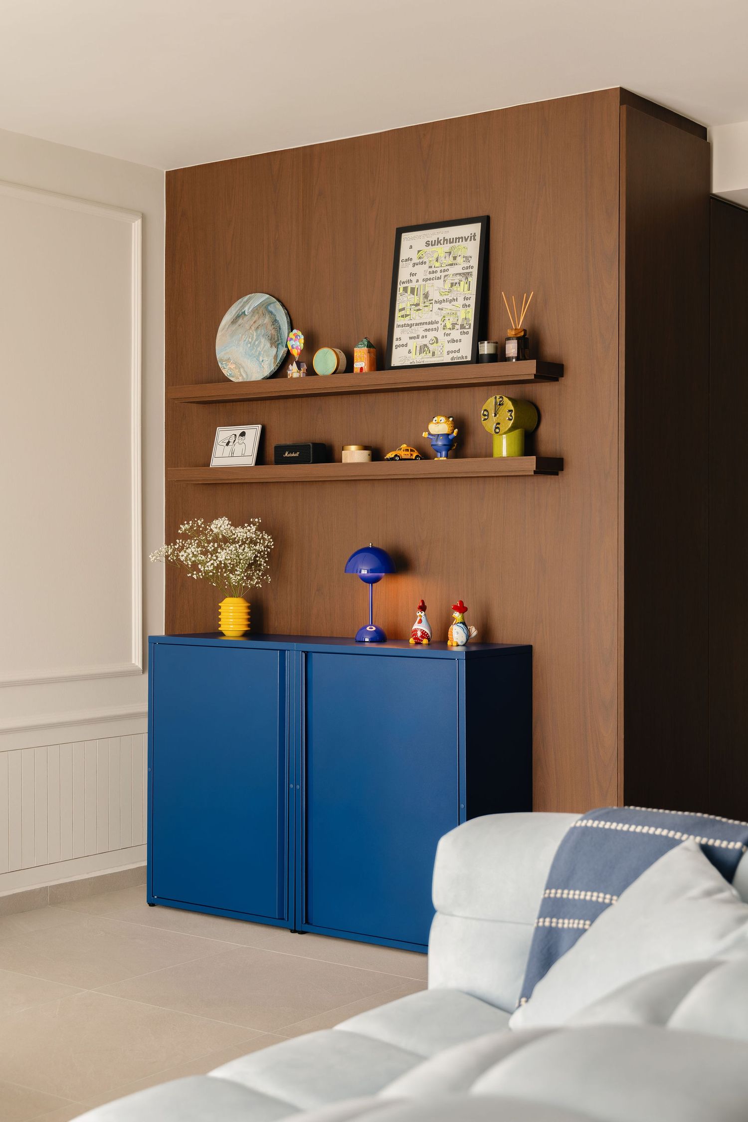 Modern living room with blue cabinet, wood-paneled wall, floating shelves with decor, and a white sofa in the foreground.