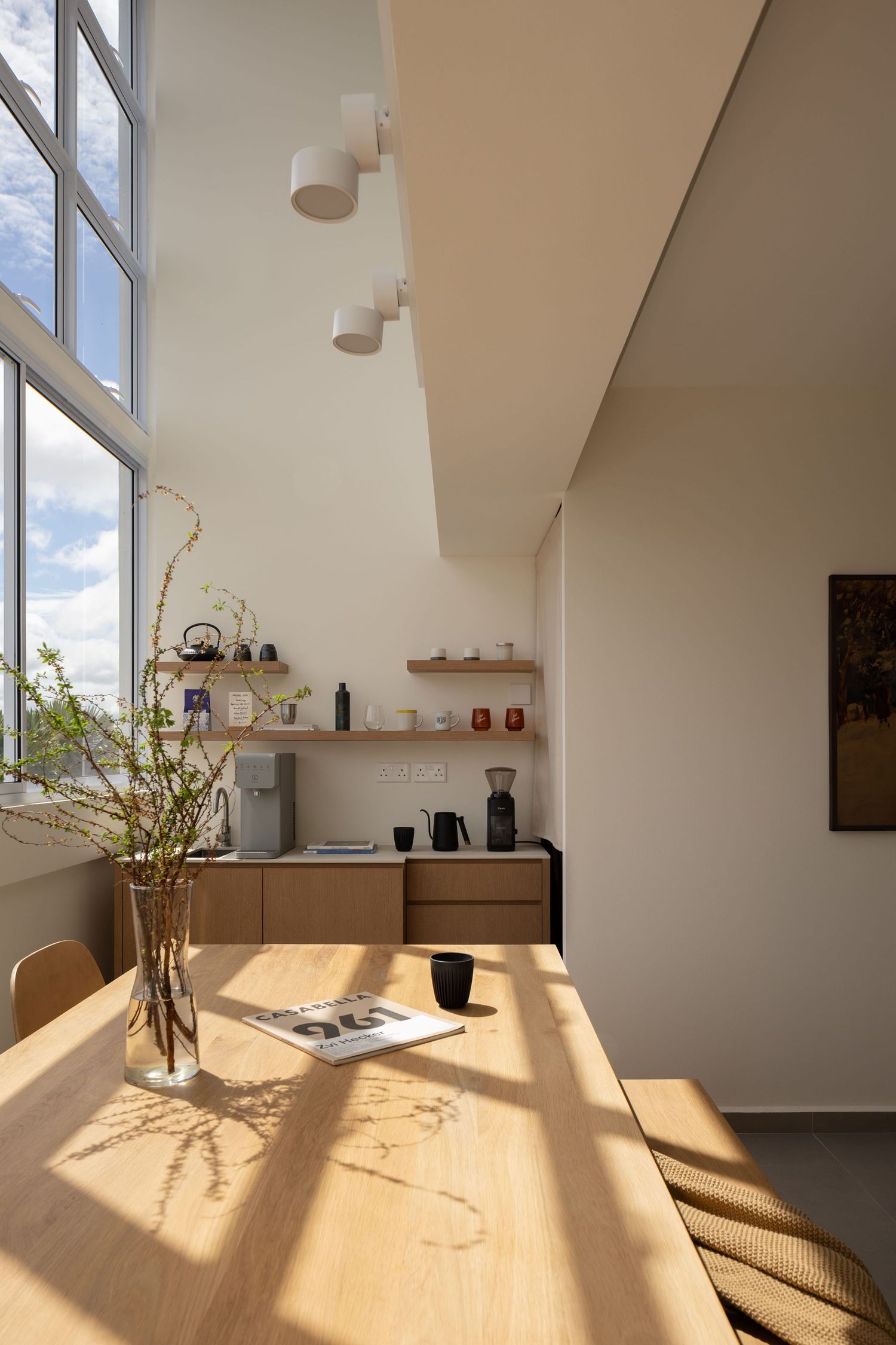 Sunlight streams across a minimalist dining area with wooden table and vase of branches in a bright modern space.