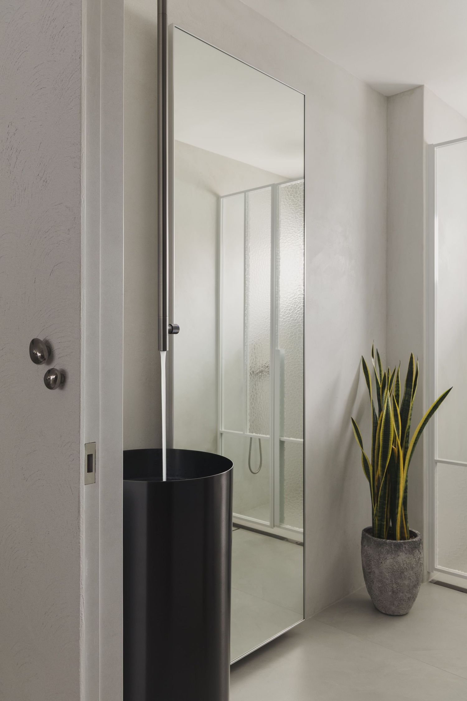 Modern minimalist bathroom hallway with large mirror, black bin, and snake plant in gray pot.