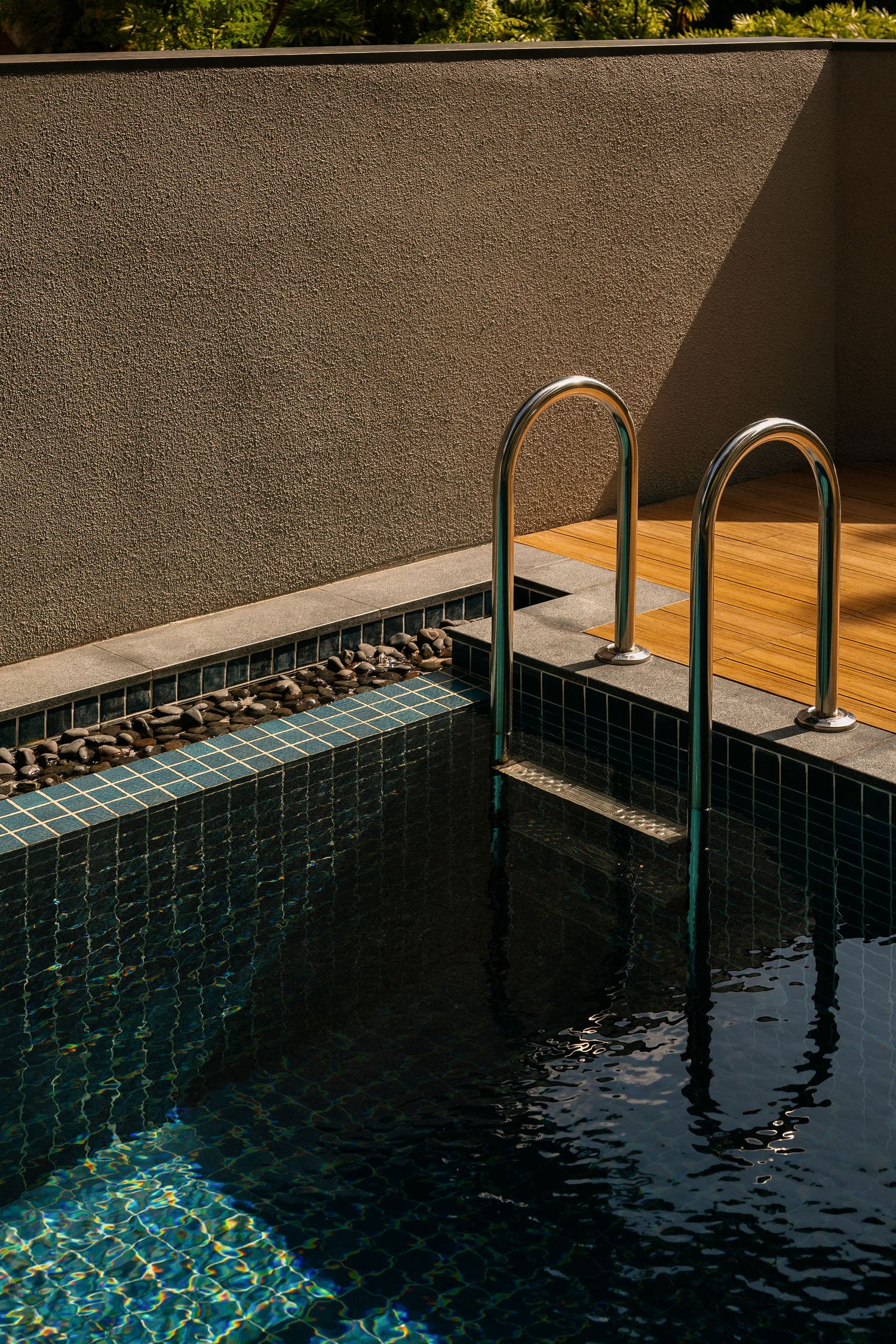 Stainless steel pool ladder rails beside dark green mosaic-tiled swimming pool with pebble border, wood deck and textured wall.