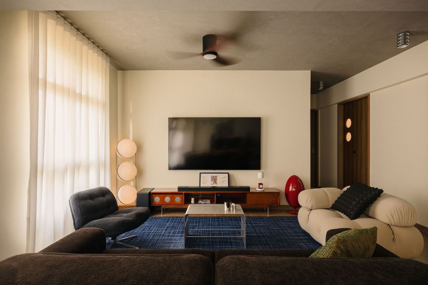 Mid-century modern living room with a wall-mounted TV, blue patterned rug, plush gray sofas, globe floor lamps, and sheer curtains.