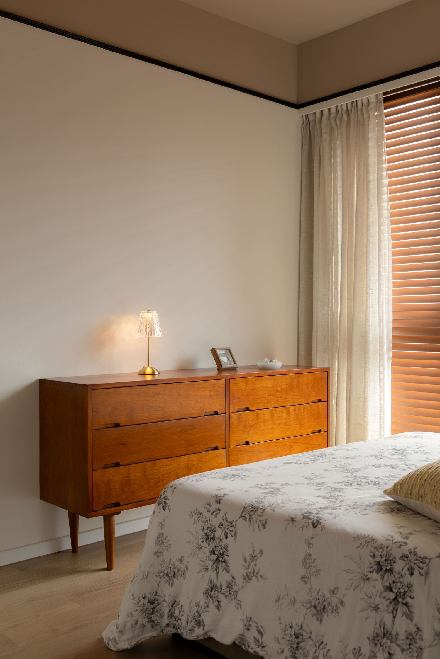 Mid-century modern bedroom with wooden dresser, brass lamp, floral bedding, and warm natural light through venetian blinds.