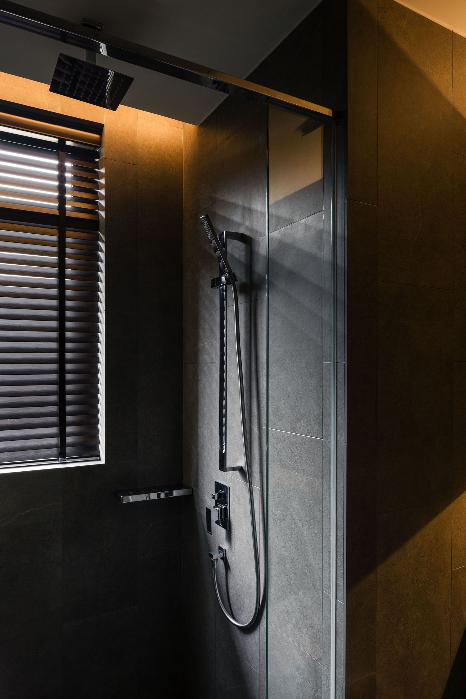 Modern black shower with rainfall head, handheld fixture, glass door, dark tiles, and window with blinds.