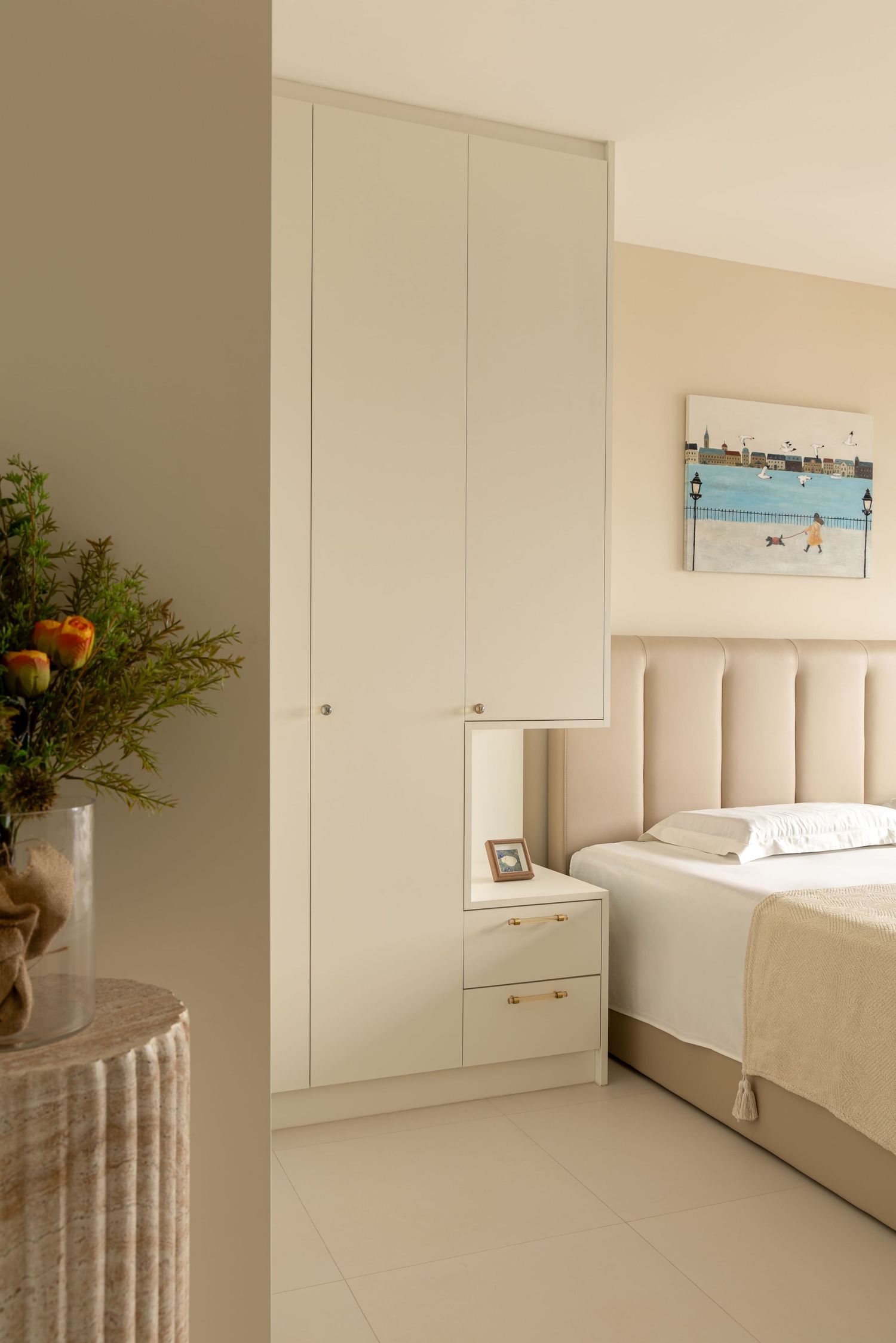 Elegant beige bedroom with tall built-in wardrobe, upholstered headboard, coastal artwork, and floral arrangement on ribbed side table.