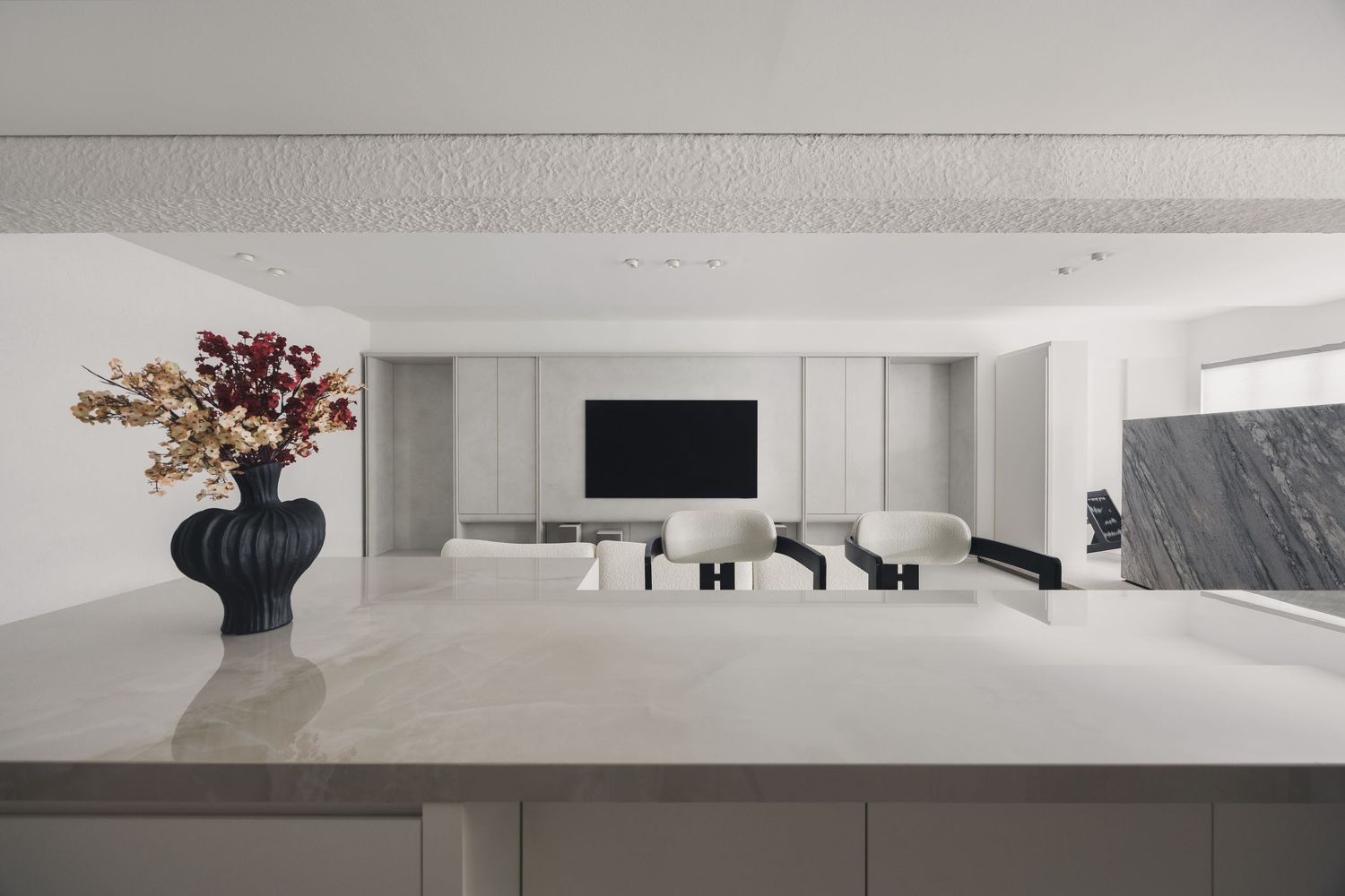 Minimalist open-plan living room with white marble surfaces, cream armchairs, wall-mounted TV, and a black vase with autumn branches.