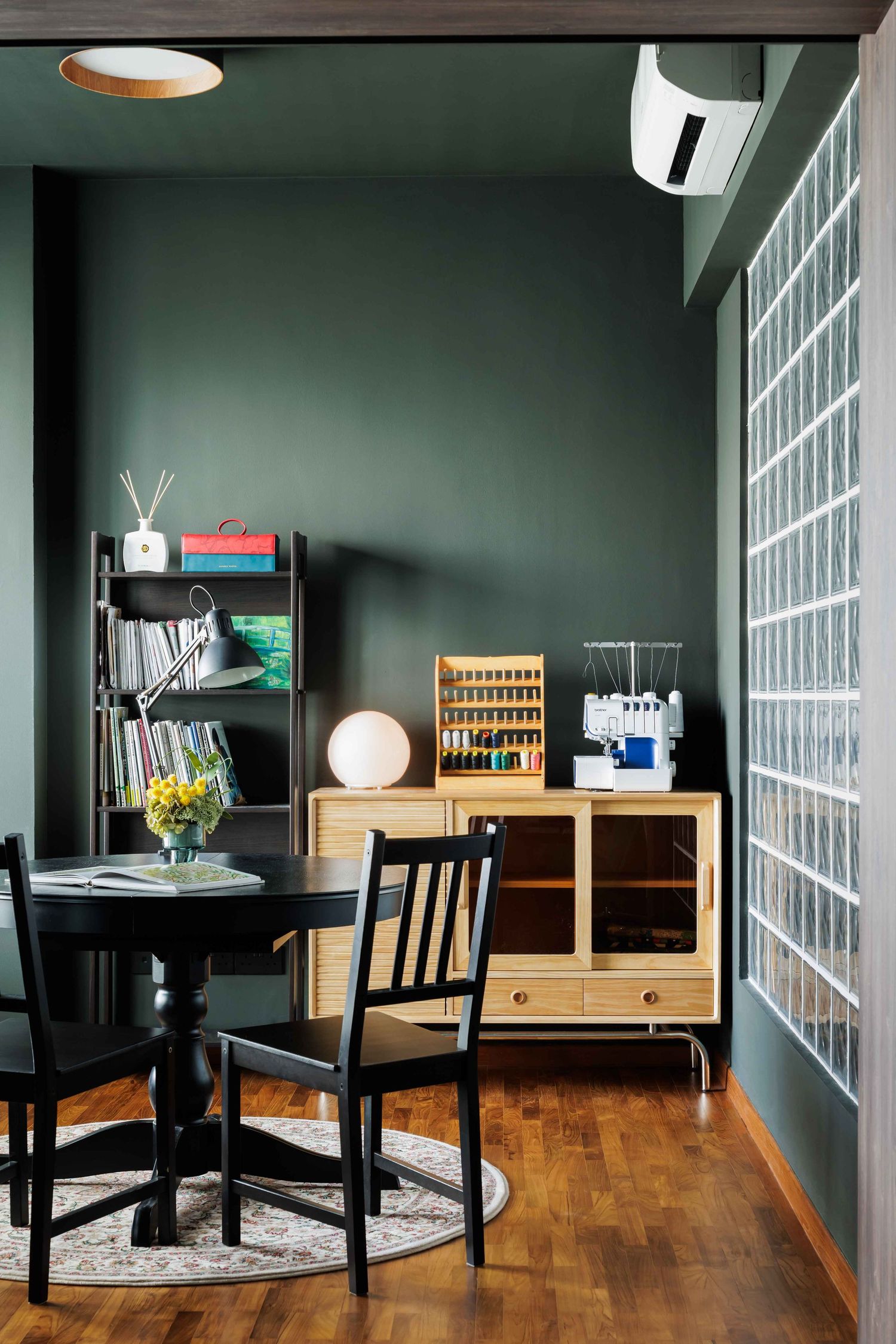 Modern home office corner with dark green walls, wood flooring, round black dining table and glass block window feature.