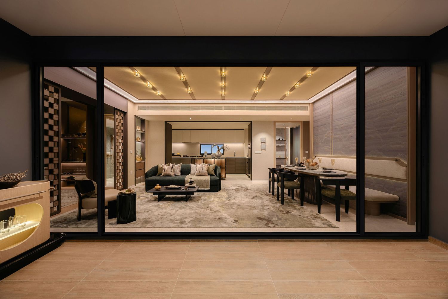 Luxury open-plan living room with plush sofas, dining table and recessed gold ceiling lights, viewed through glass doors at night.