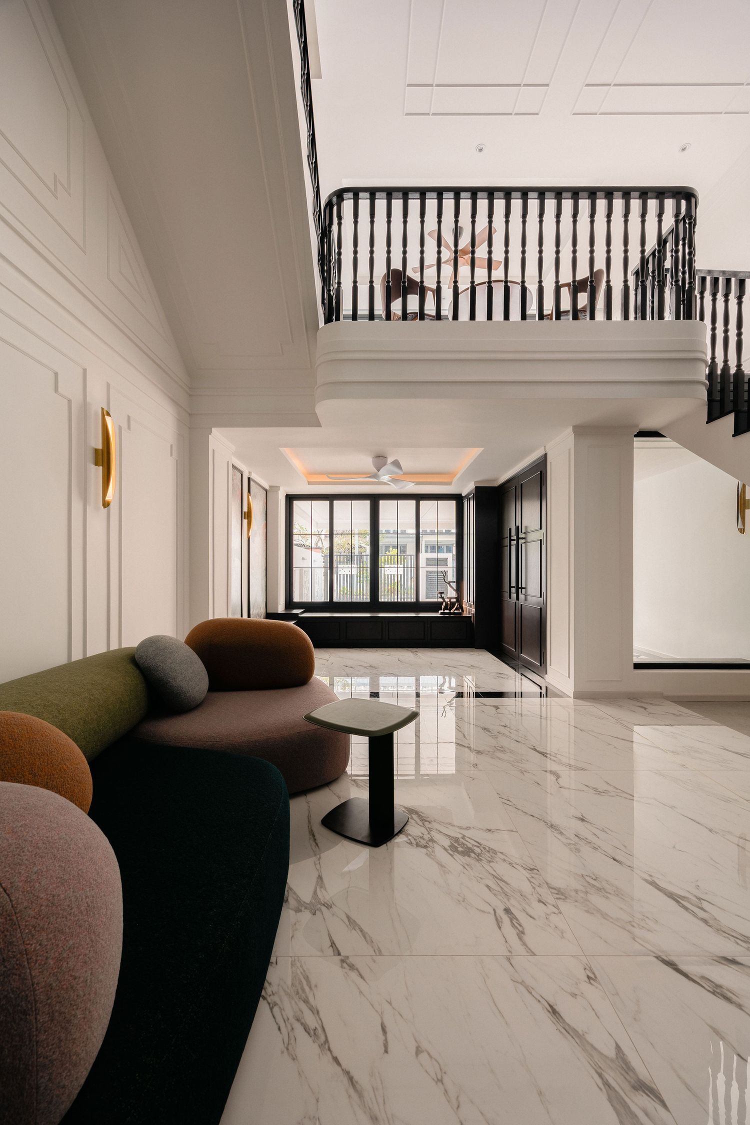 Elegant double-height living room with white marble floors, curved multicolor sofas, black balustrade mezzanine and classic wall paneling.