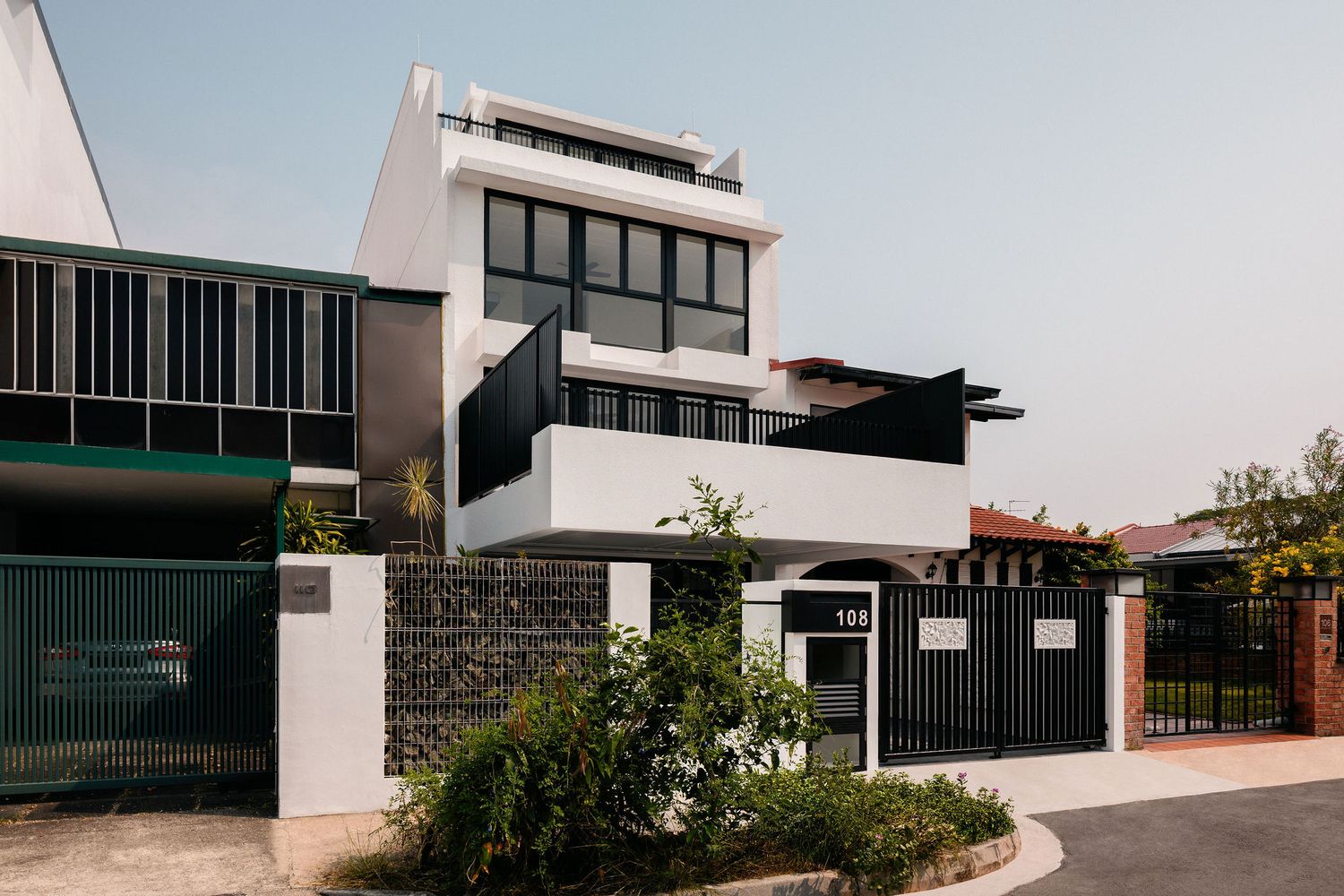 Modern white three-story house with black-framed windows, a rooftop terrace, stone gabion wall and black gate on a quiet residential street.