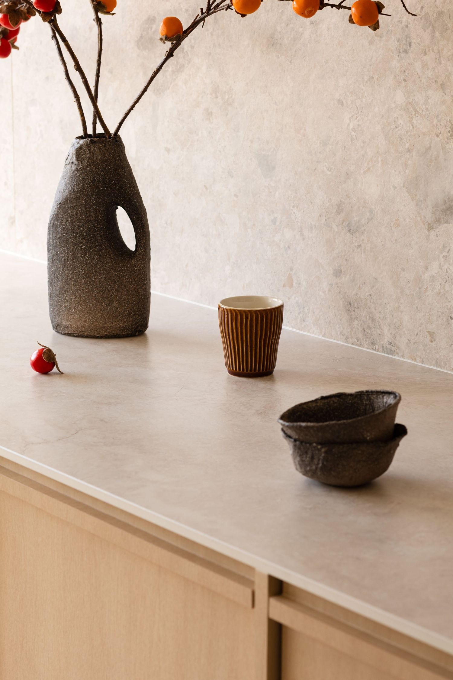 A minimalist still life composition with orange and red flowers in a gray ceramic vase on a light-colored surface.