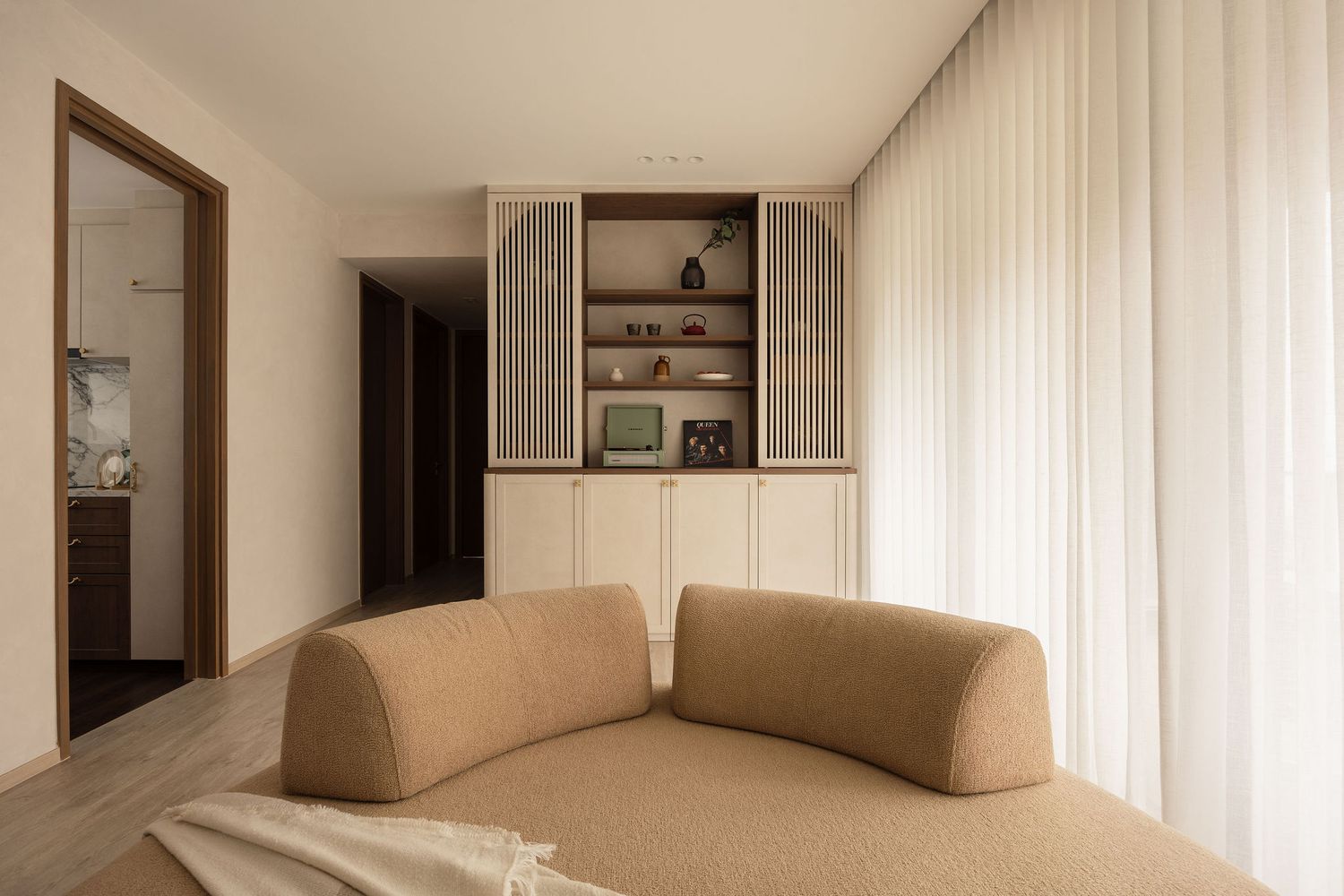 Modern living room with curved beige sofa, built-in shelving unit, and sheer white curtains in a neutral toned interior.