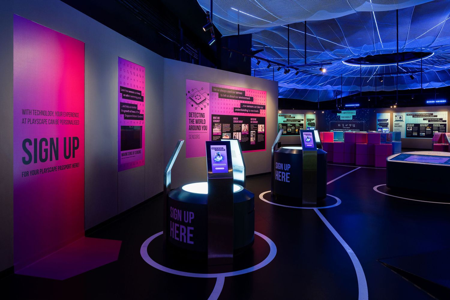 Interactive museum exhibit with neon pink and purple lighting, sign-up kiosks, and engaging displays on dark flooring.