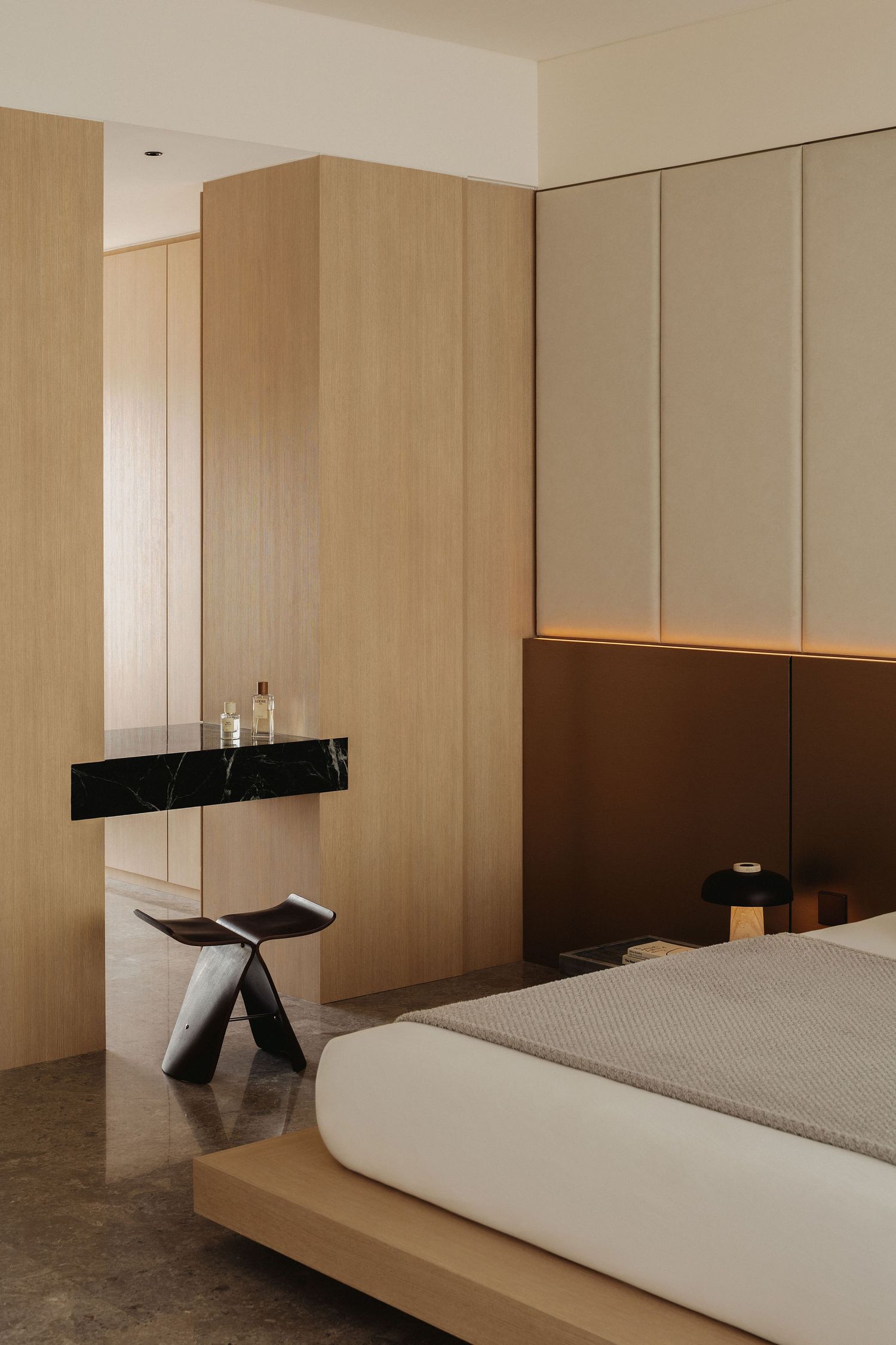 Minimalist bedroom with light wood wardrobes, black marble vanity, sculptural stool, and upholstered bed with warm accent lighting.