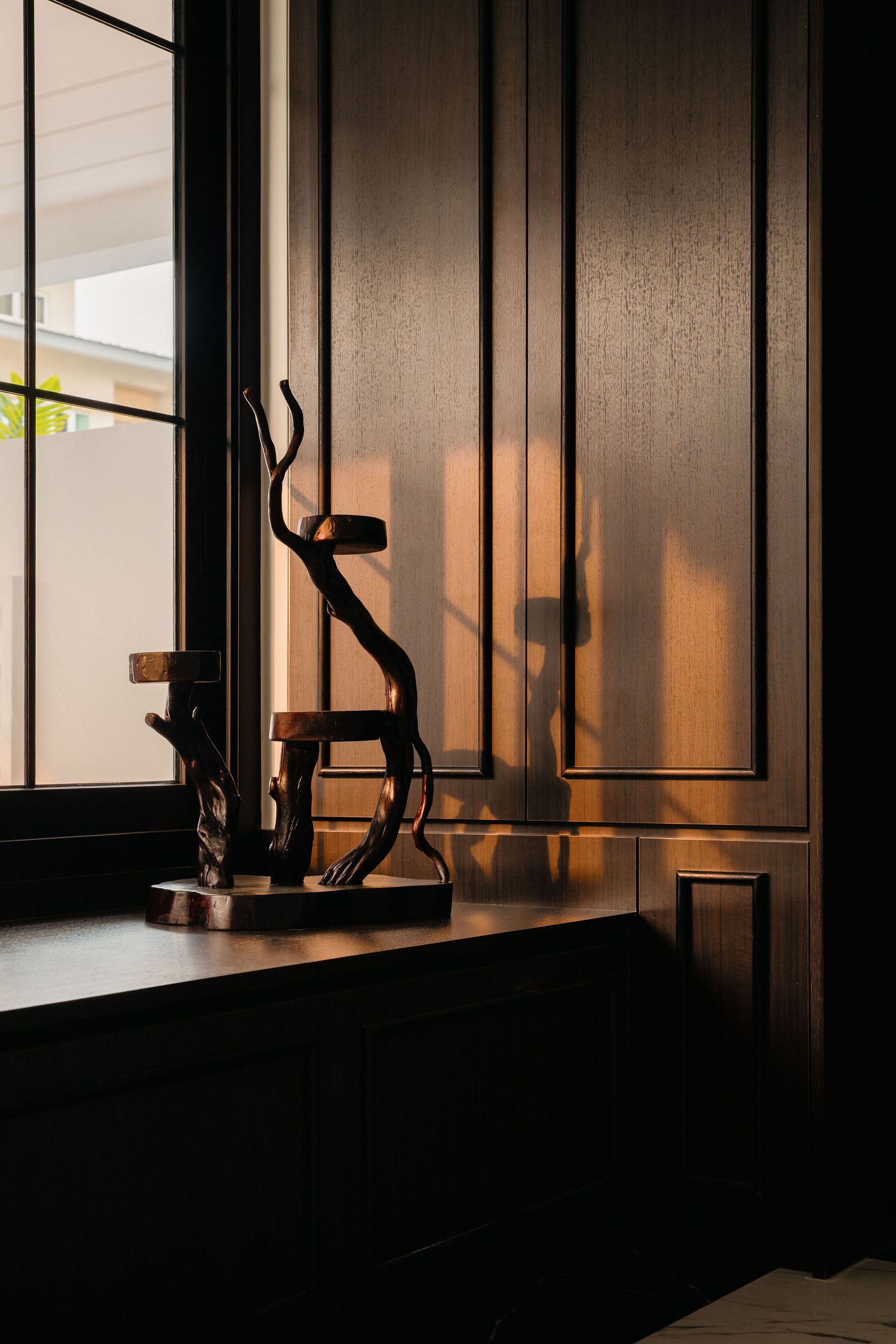 Sculptural driftwood candle holder on a dark wood windowsill, lit by warm sunlight beside elegant paneled cabinetry.
