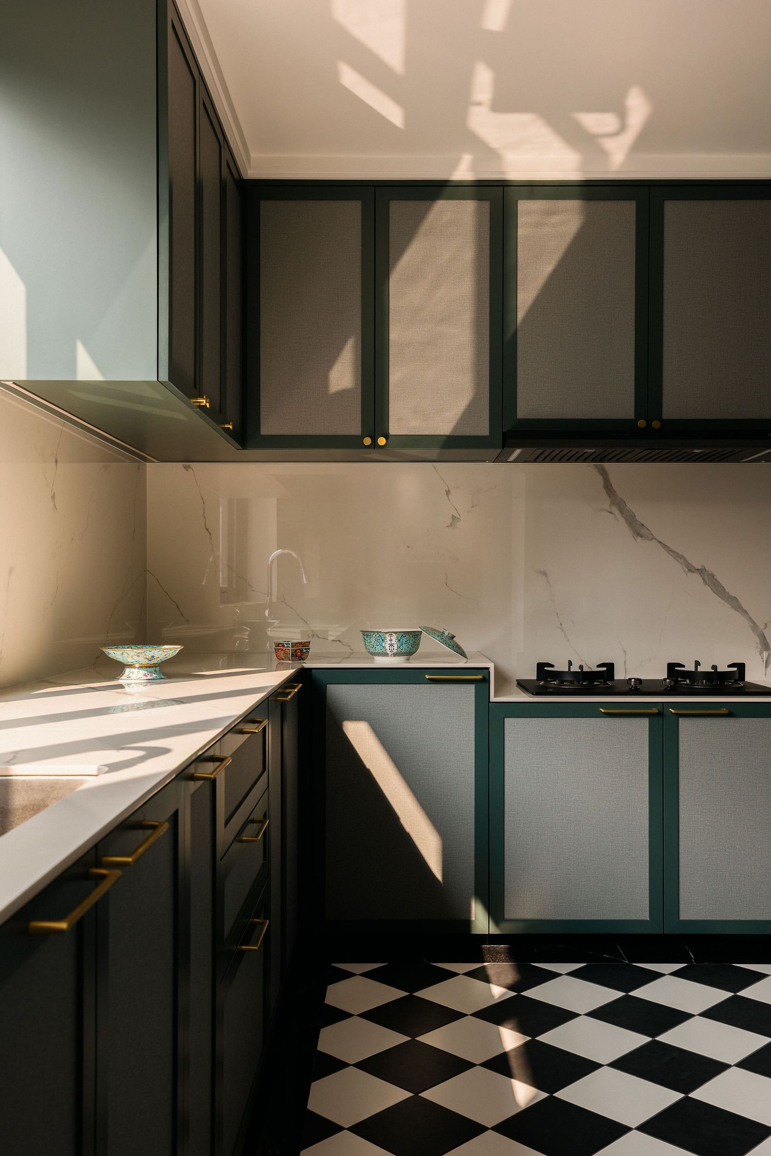 Modern L-shaped kitchen with dark green cabinets, brass handles, white marble backsplash and a black and white checkered tile floor.