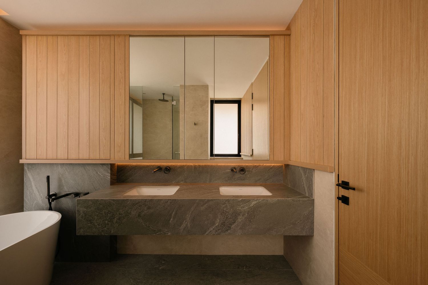 Modern bathroom with light wood paneling, a double vanity on a gray stone countertop, mirrored cabinet, and a freestanding white bathtub.