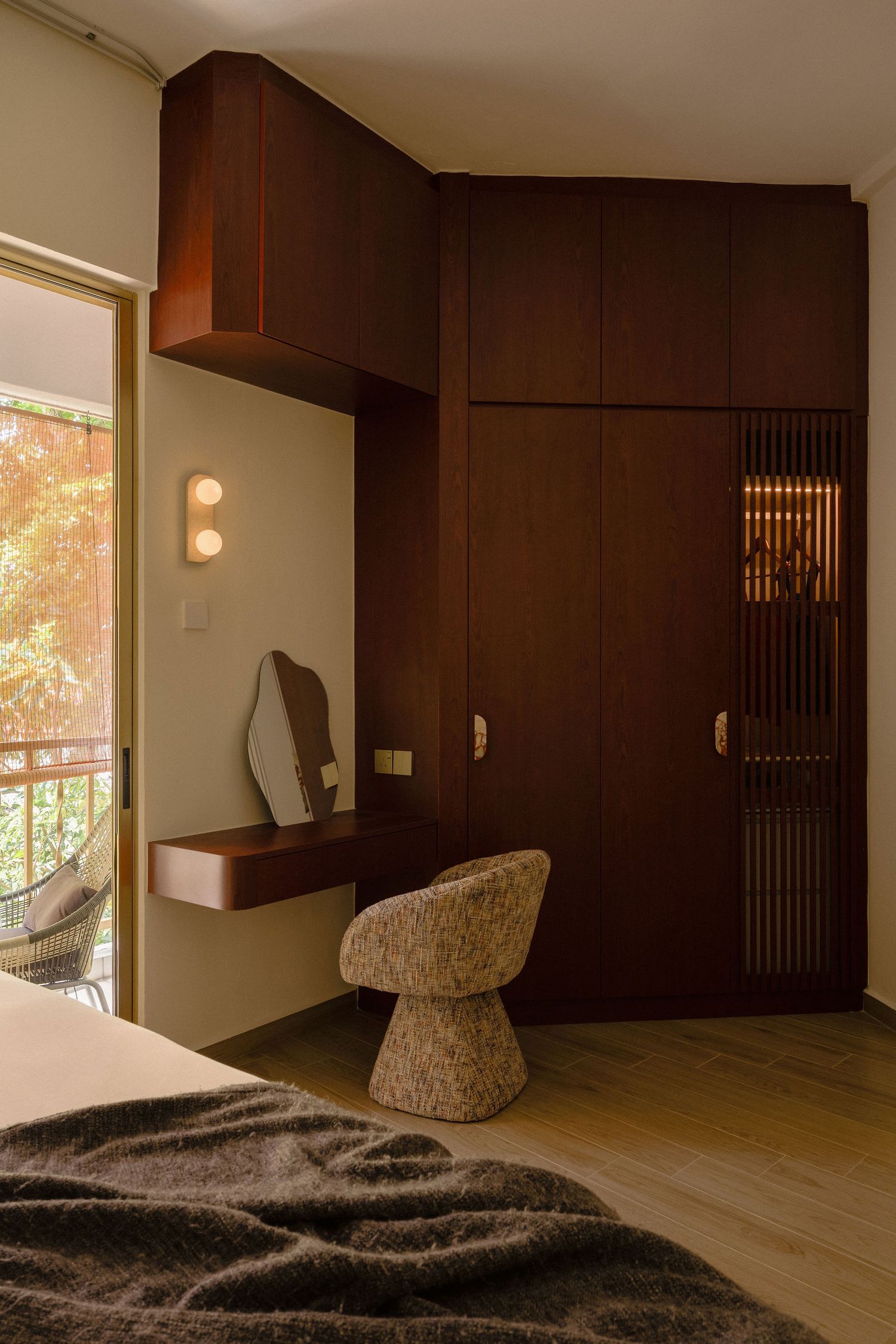 Modern bedroom corner with dark walnut built-in wardrobe, floating vanity desk, organic mirror, textured accent chair and balcony door.
