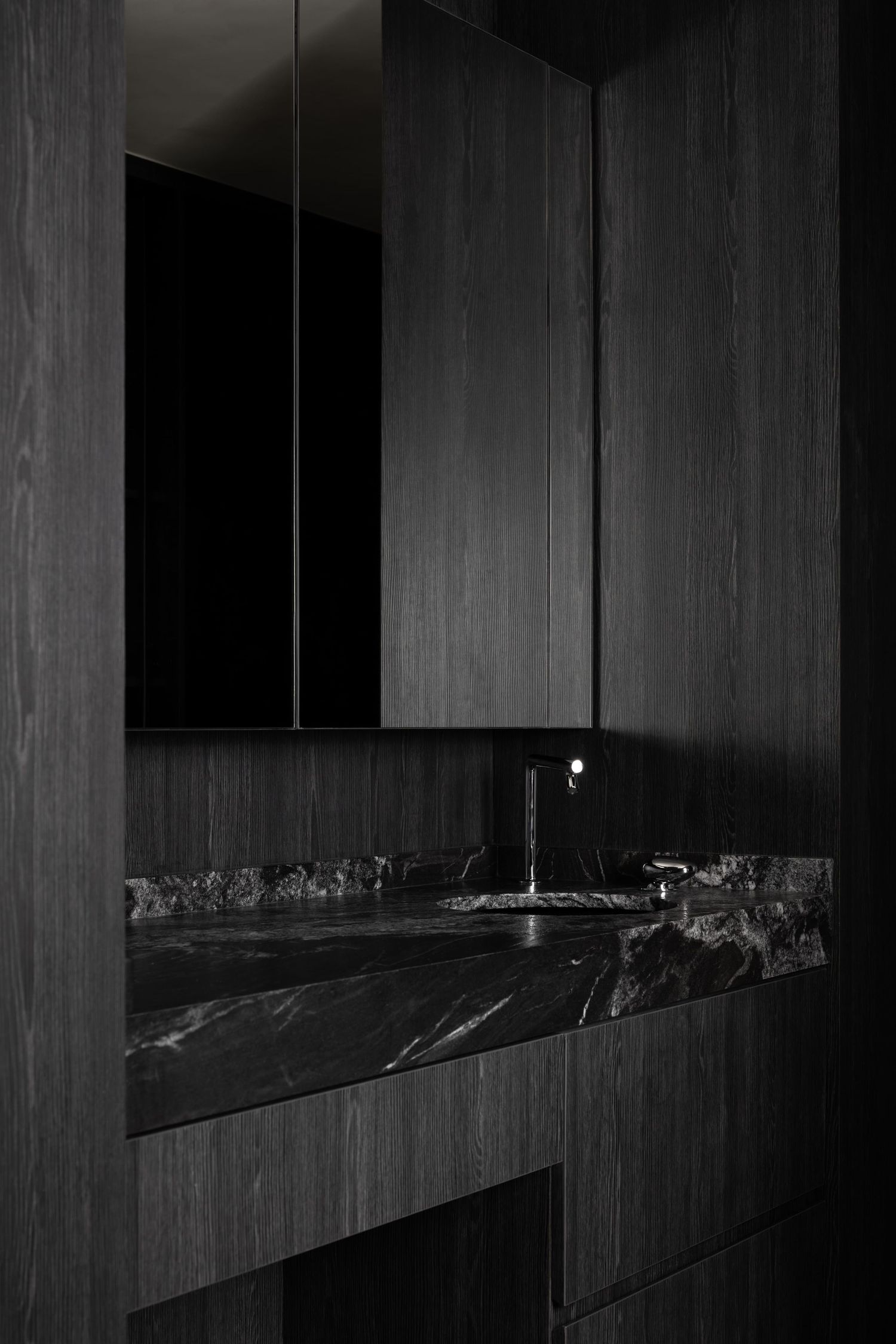 Modern black marble bathroom vanity with minimalist faucet and wooden wall panels in dark moody lighting.
