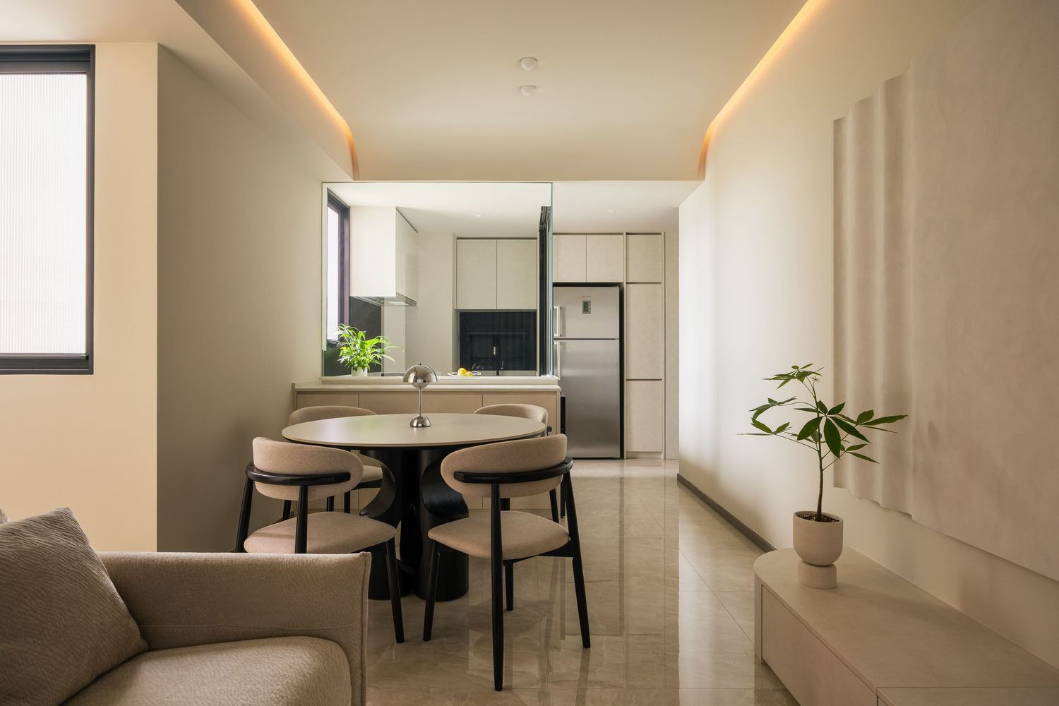 Modern open-plan dining area with round table, beige chairs, and sleek kitchen featuring stainless steel refrigerator.