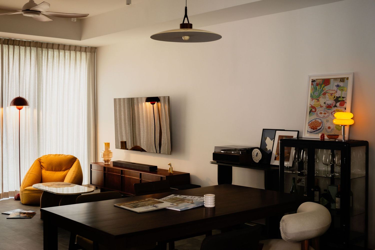 Modern living room with dark dining table, yellow armchair, wall-mounted TV, pendant light, and decorative cabinet.