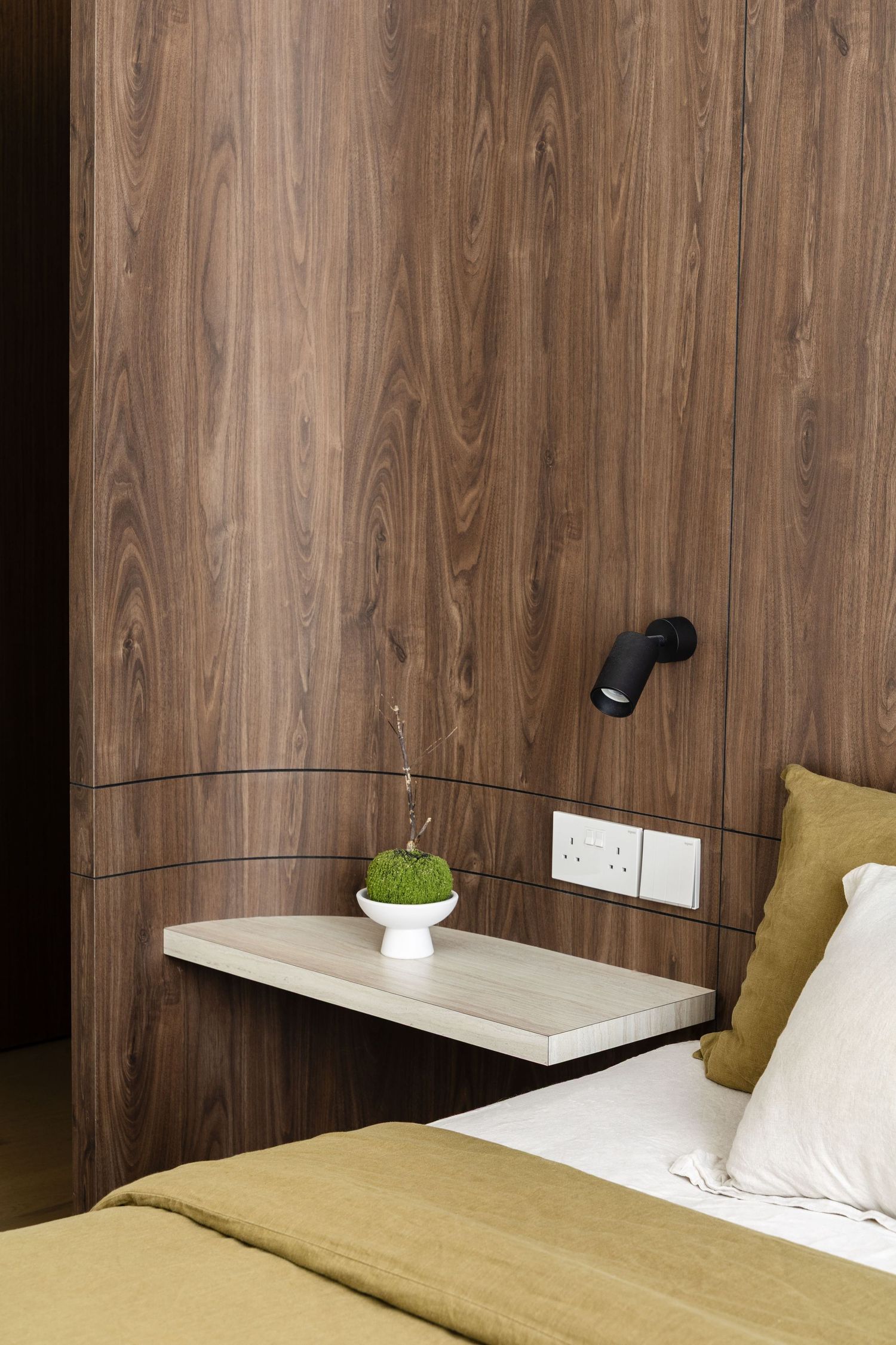 Black adjustable wall sconce light fixture mounted on wood paneled wall above modern minimalist bed with yellow cushions.