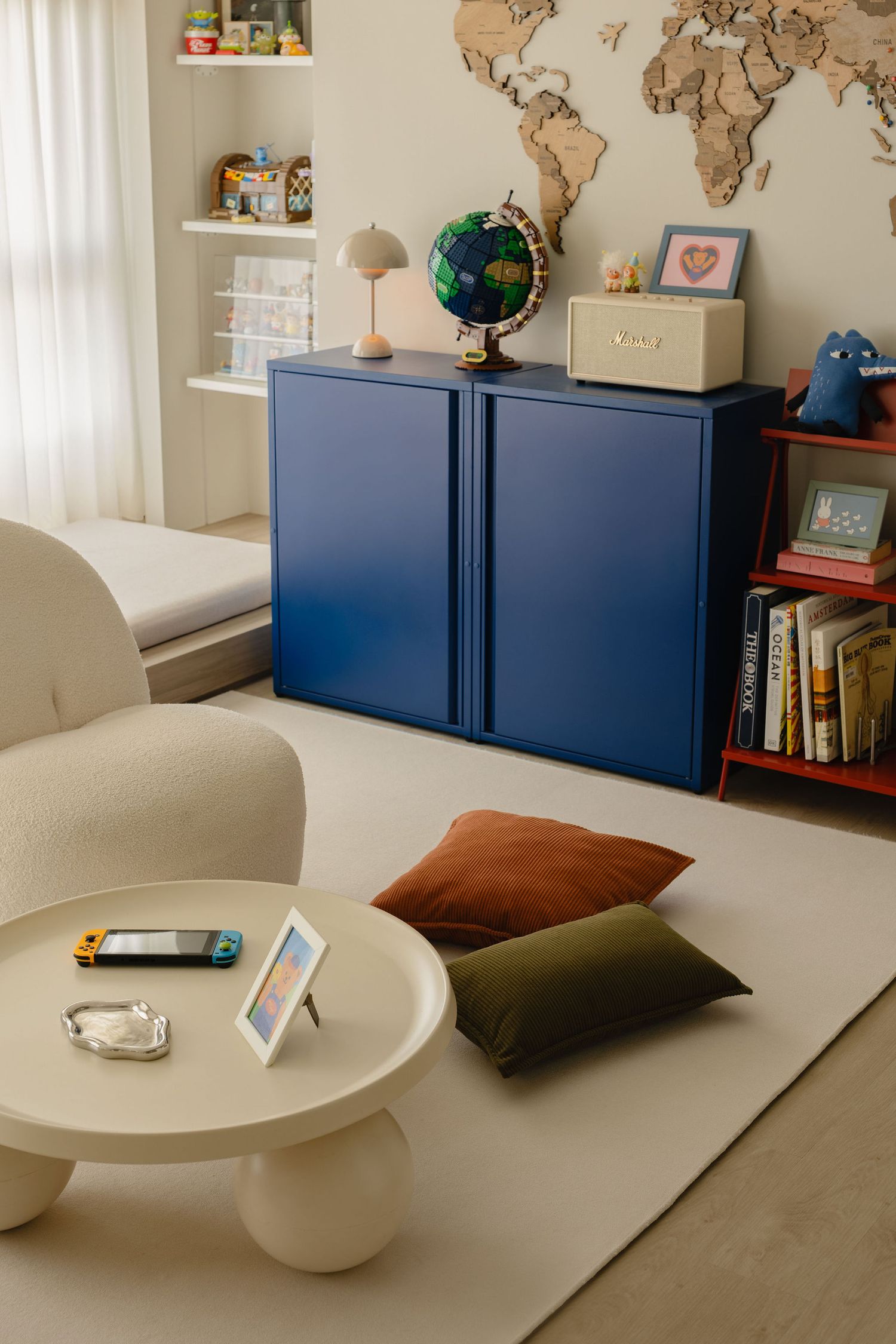 Modern kids room with blue cabinet, globe, world map wall art, red bookshelf, round white coffee table, and colorful pillows.