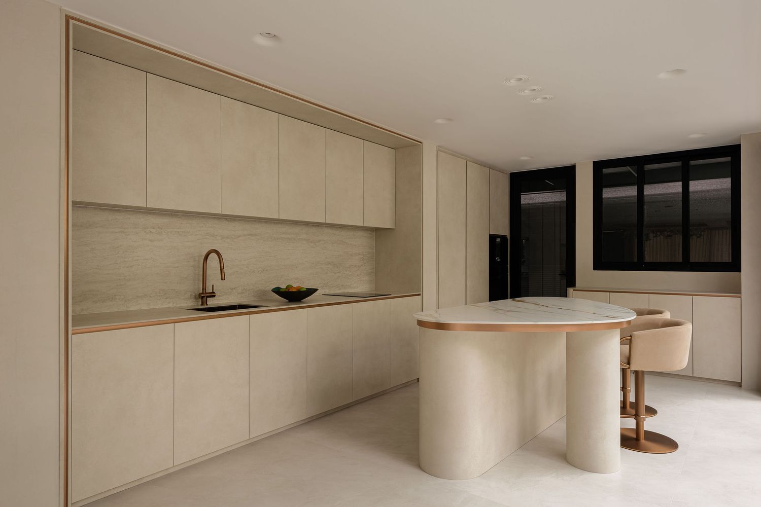Minimalist kitchen with beige cabinetry, curved island, bar stools, brass sink fixture, and large black-framed windows.