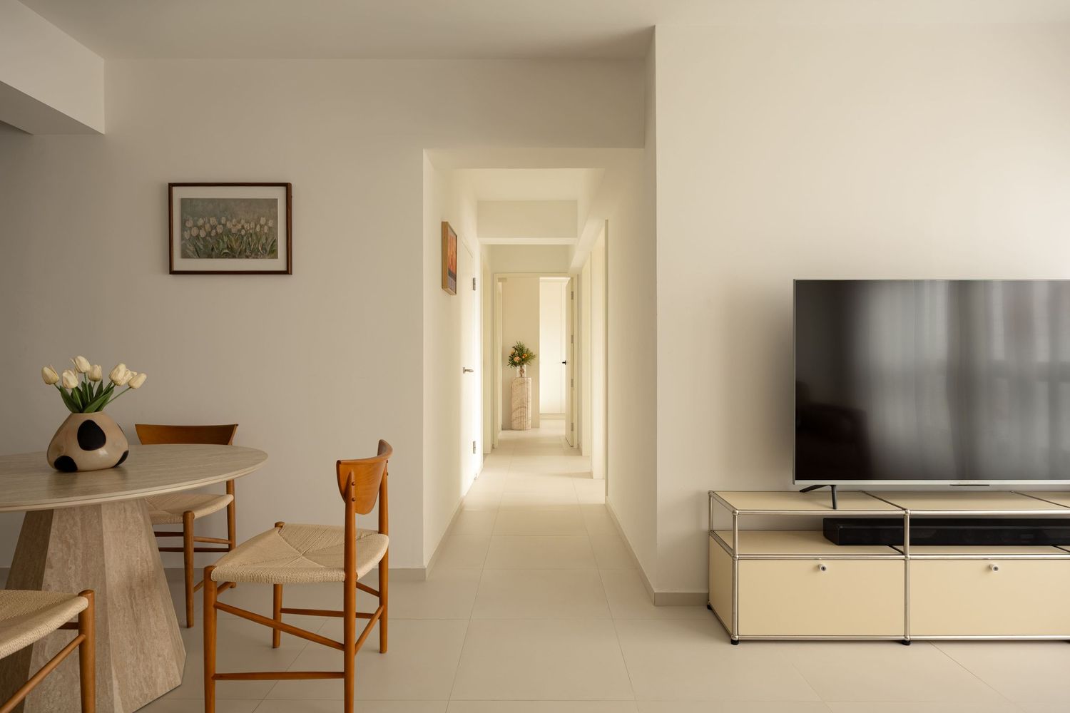 Minimalist living room with TV on metal stand, wooden dining table, chairs, and bright hallway in modern apartment.
